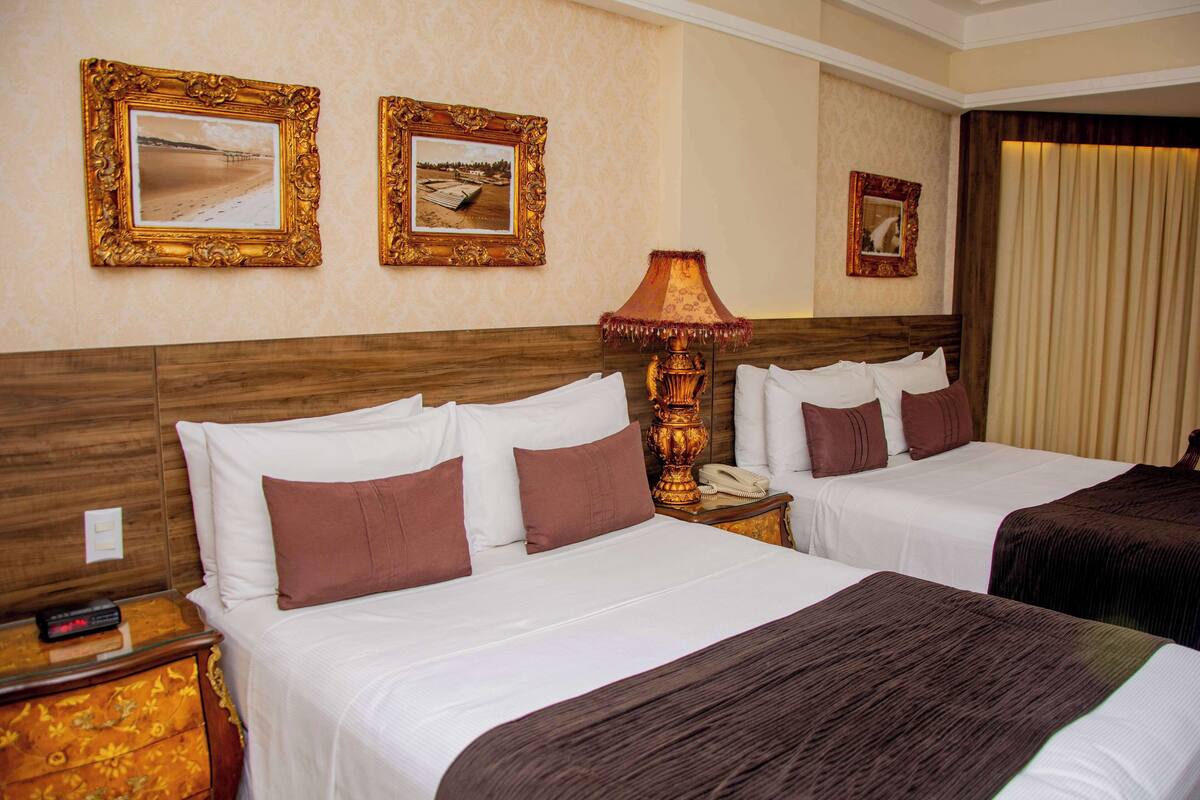 A hotel room with two beds, a wooden headboard, a nightstand with a lamp, and framed pictures on the wall.