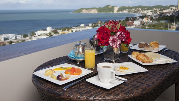 Ein Frühstückstisch mit Fruchtplatten, Gebäck, Kaffee und Saft, mit Blick auf die Küste, Gebäude und Hügel.
