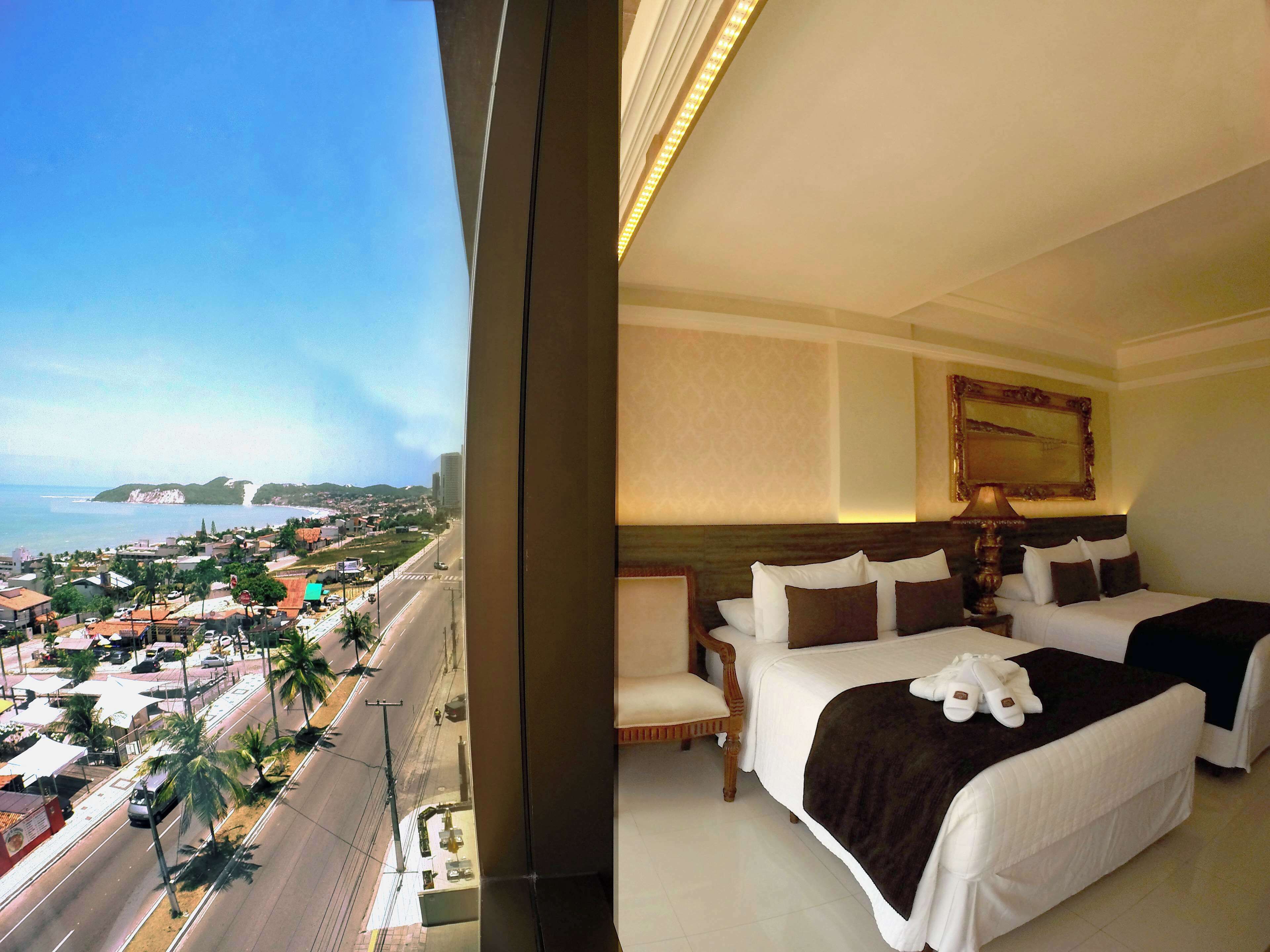 A hotel room with two beds, a view of the city and coastline, a painting on the wall, and a lamp.