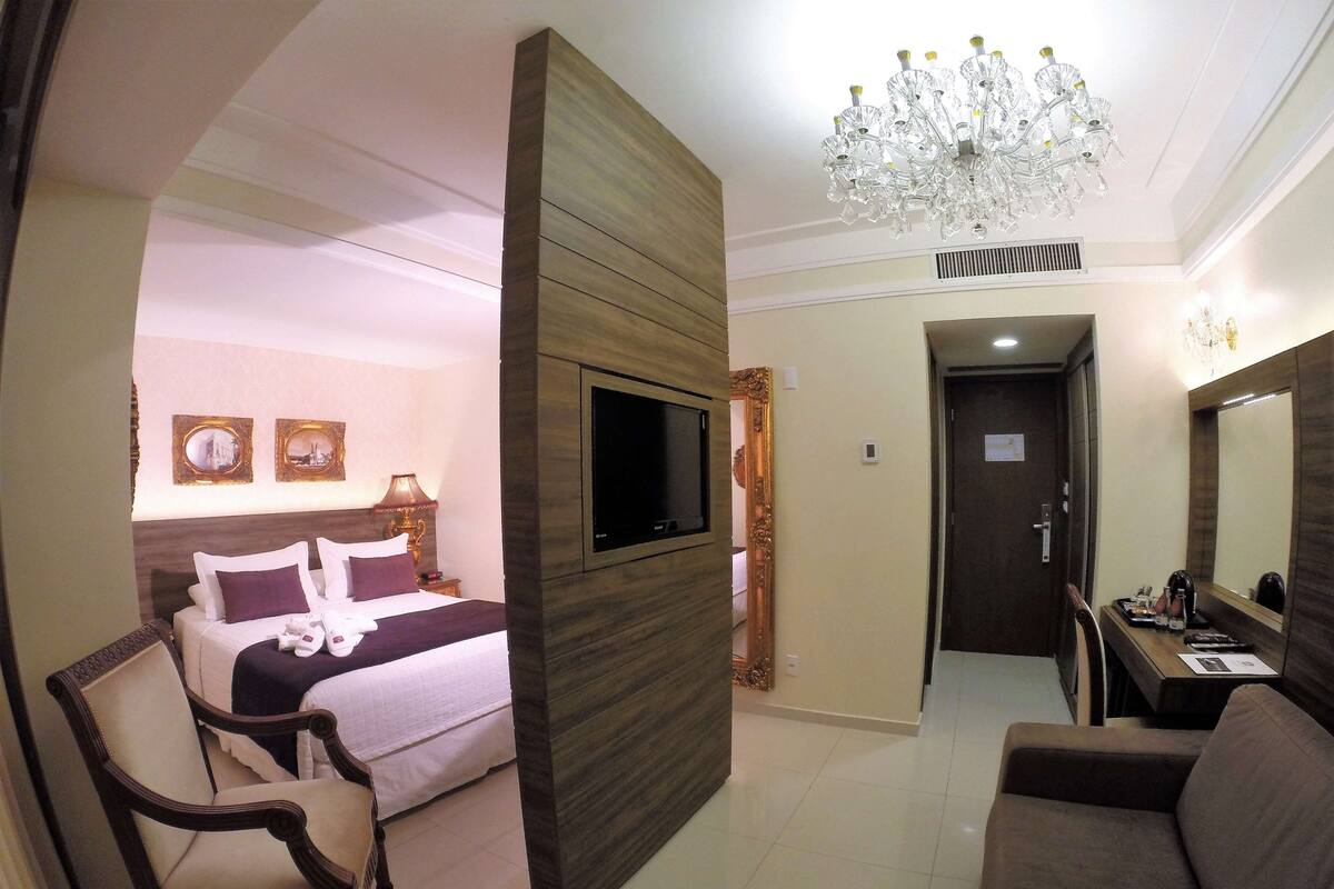 A hotel room with a bed, a TV mounted on the wall, a chair, a sofa, and a chandelier.