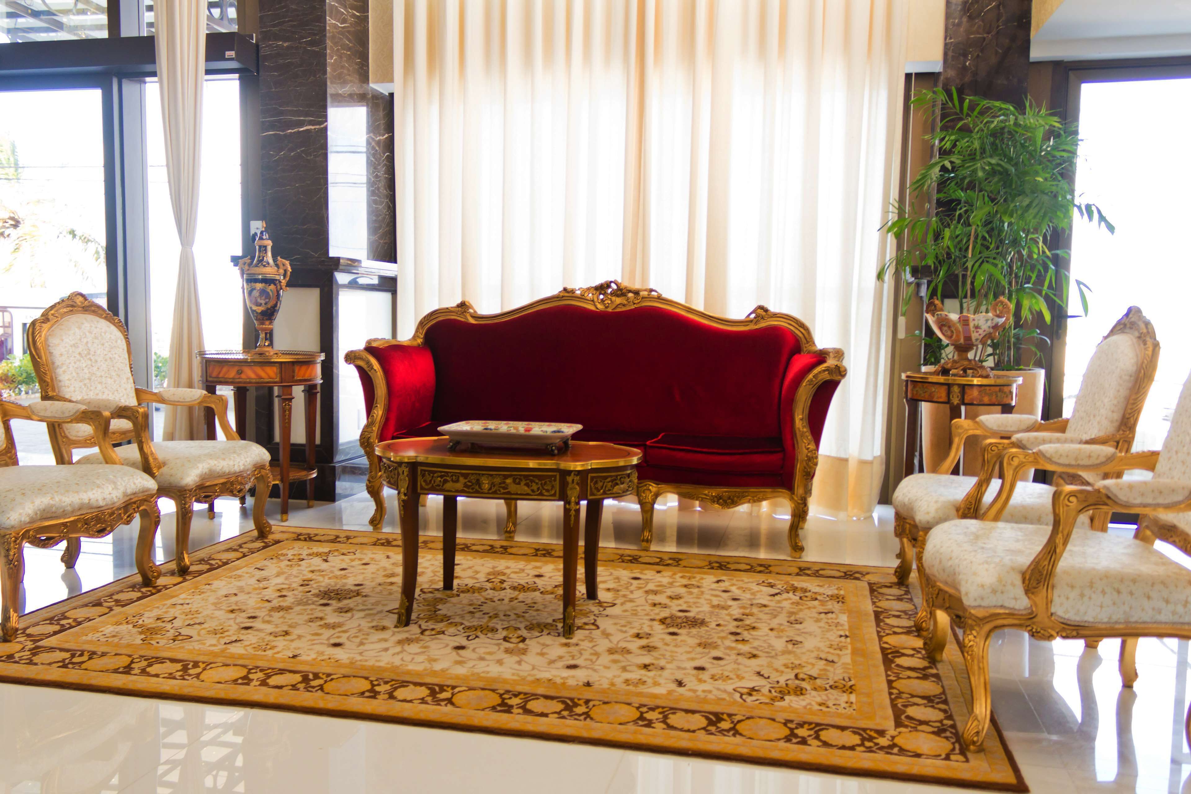 Une chambre luxueuse avec un canapé en velours rouge, des fauteuils richement décorés, un tapis à motifs et une grande plante en pot.
