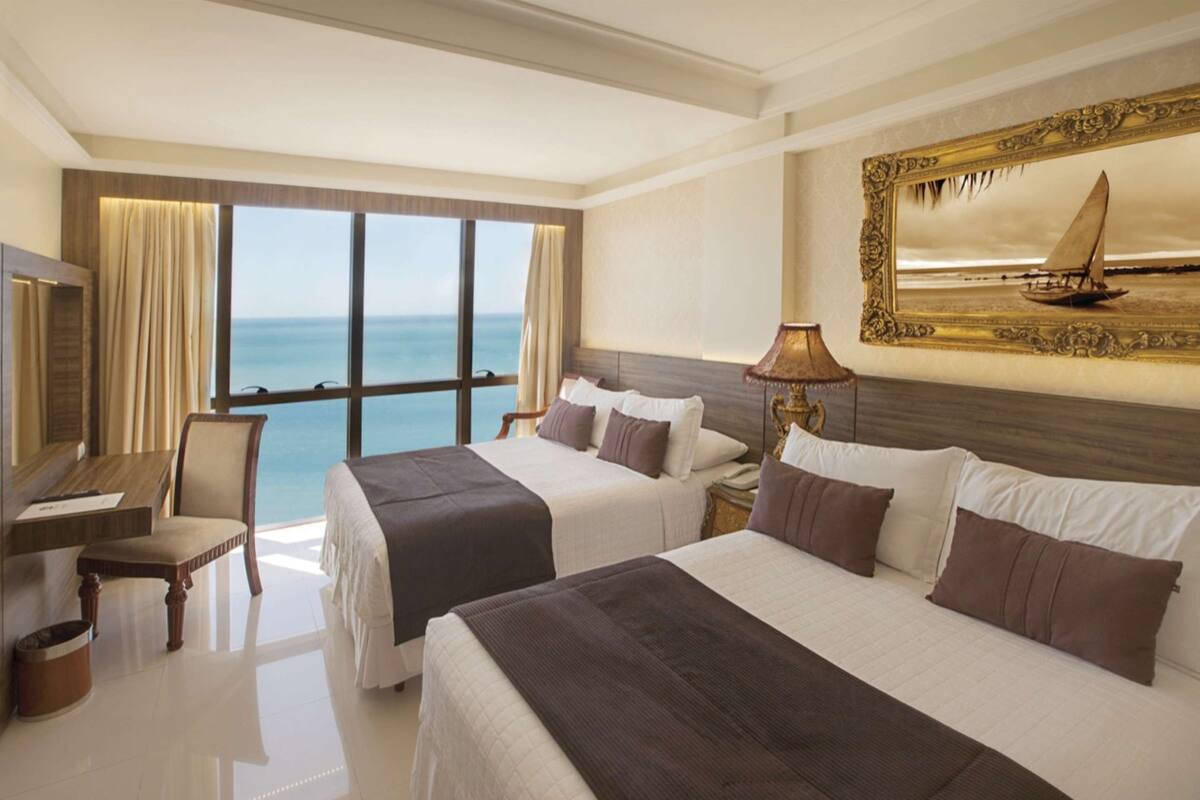 A hotel room with two beds, a desk, a chair, and a large window offering a sea view.