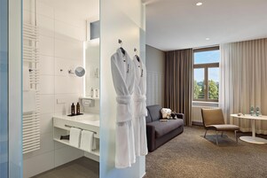 Quarto de hotel moderno com um espelho grande, toalhas brancas, um sofá, uma cadeira e uma pequena mesa com produtos de higiene pessoal.