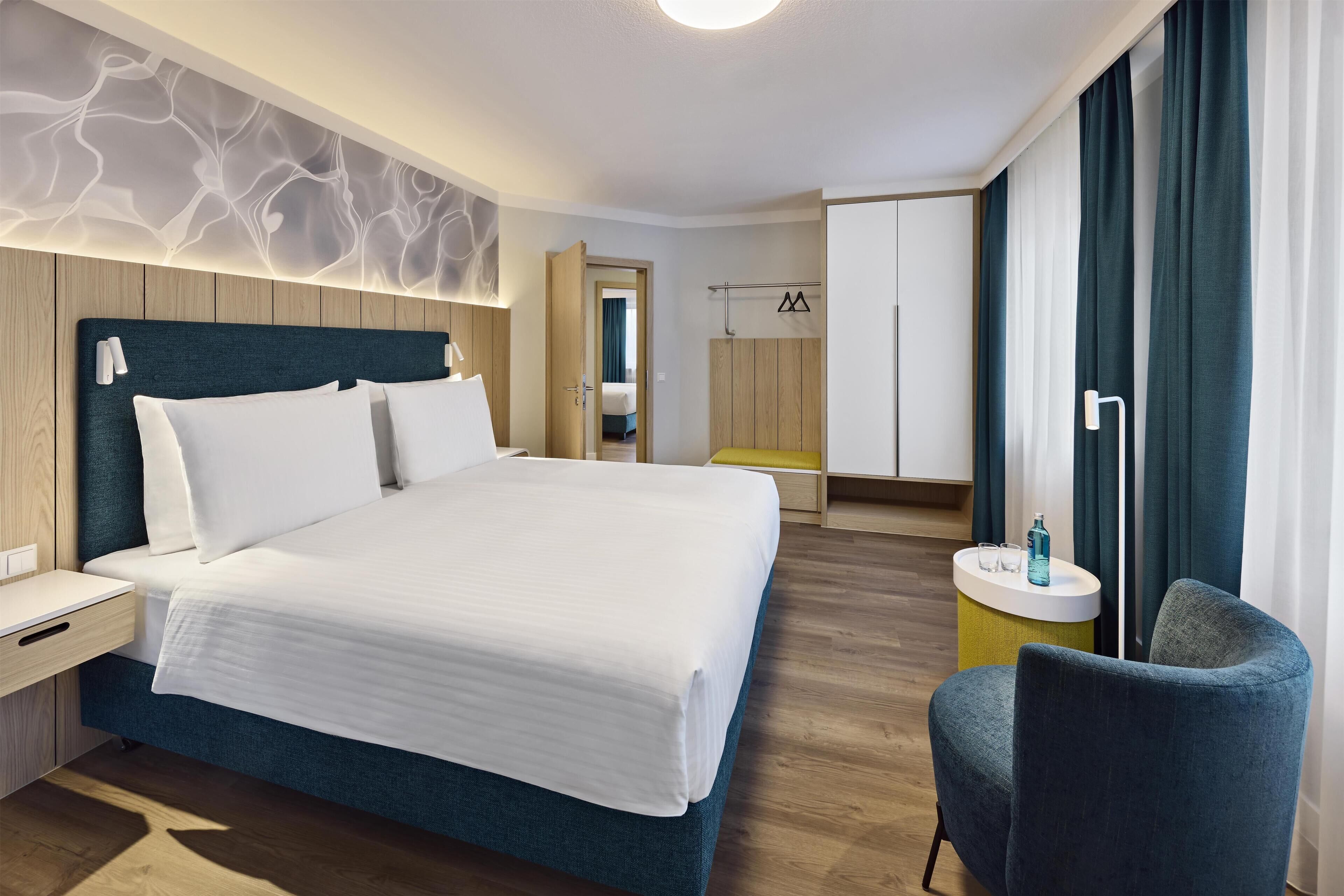A modern hotel room with a large bed, bedside tables, a chair, and a small round table with a lamp.