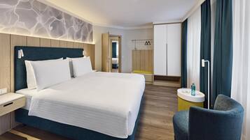 A modern hotel room with a large bed, bedside tables, a chair, and a small round table with a lamp.