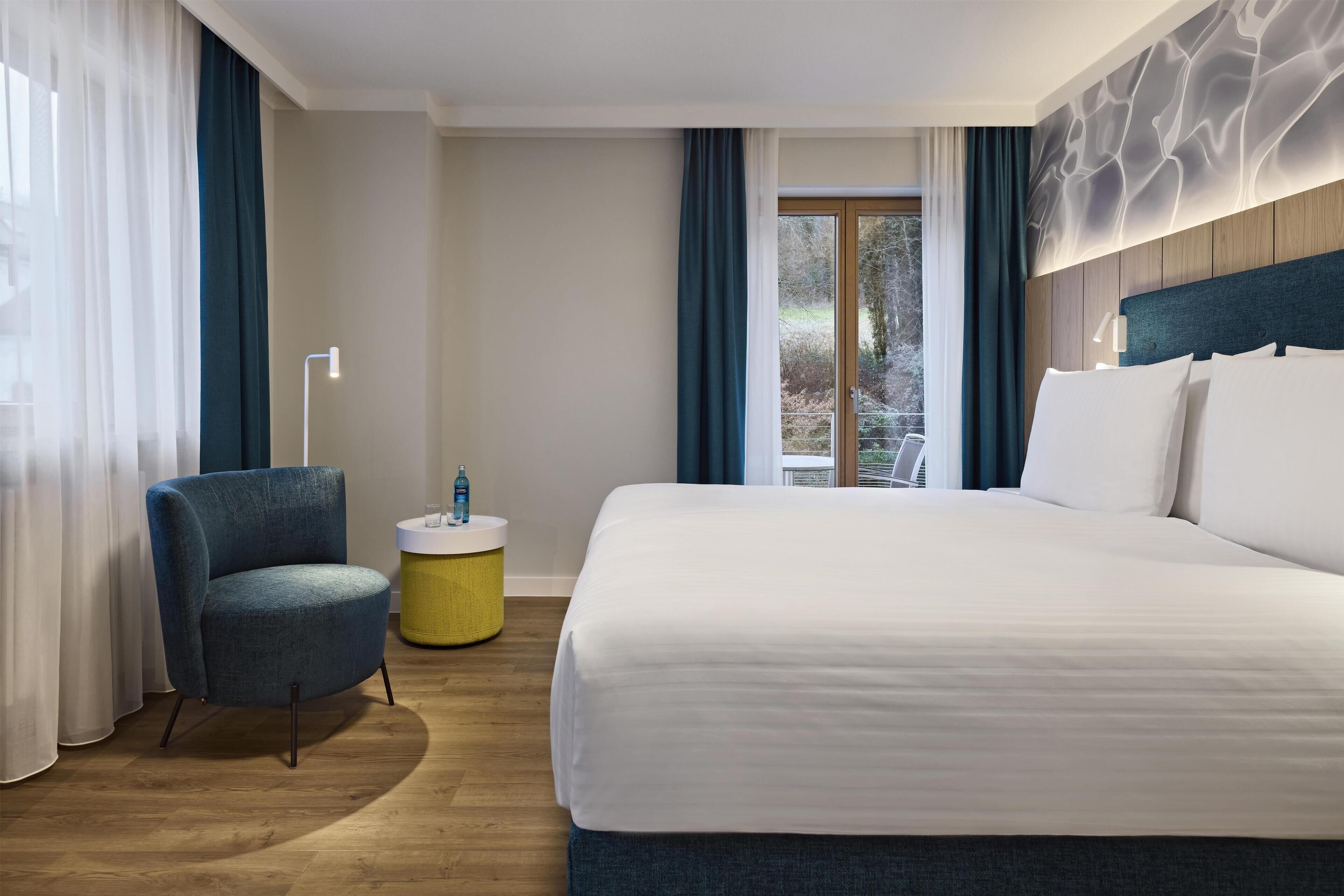 Une chambre d’hôtel avec un grand lit, une chaise, une petite table ronde et une fenêtre avec des rideaux.