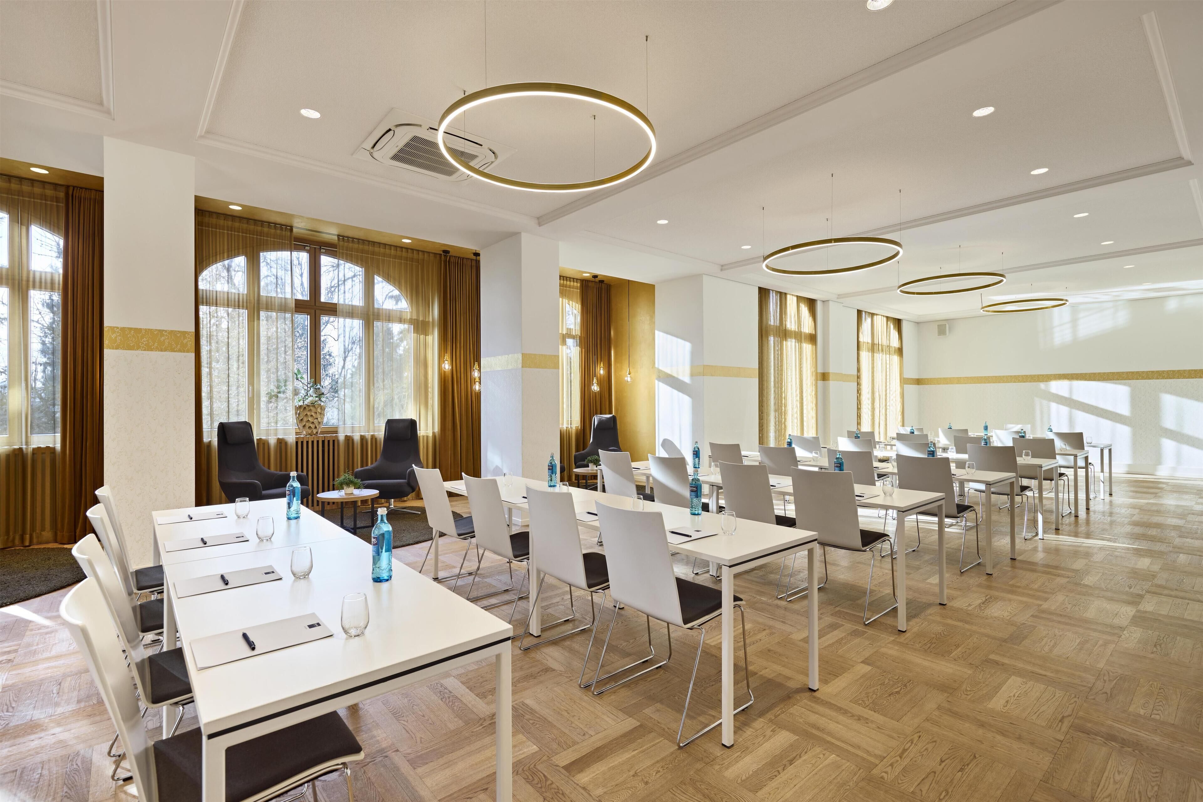 Un salón de conferencias con varias mesas y sillas, amplias ventanas con cortinas, e iluminación moderna.