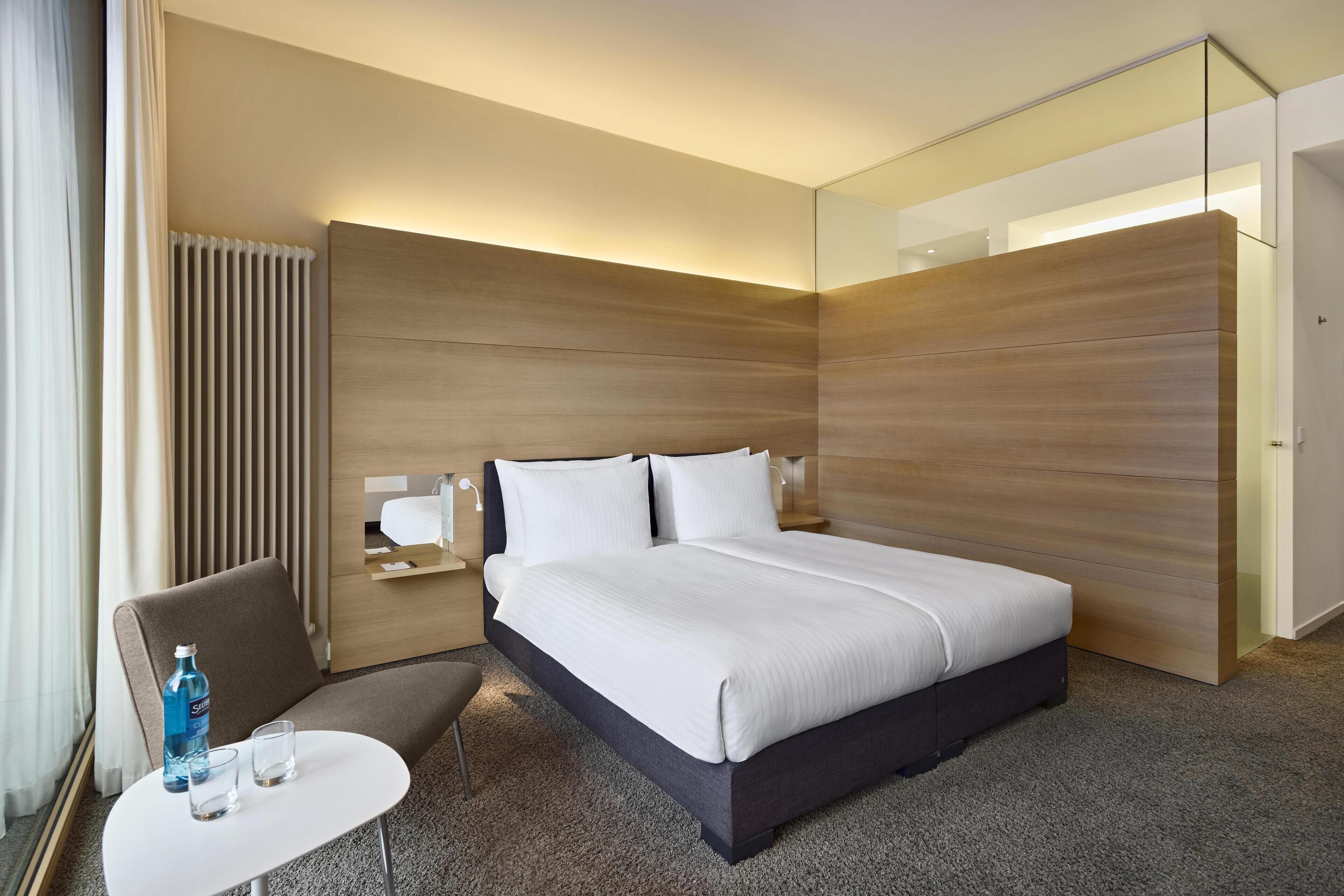 A modern hotel room with a large bed, a seating area with a table, and a wardrobe.