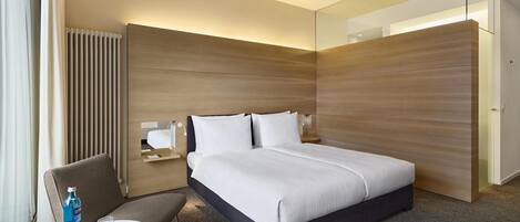 A modern hotel room with a large bed, a seating area with a table, and a wardrobe.