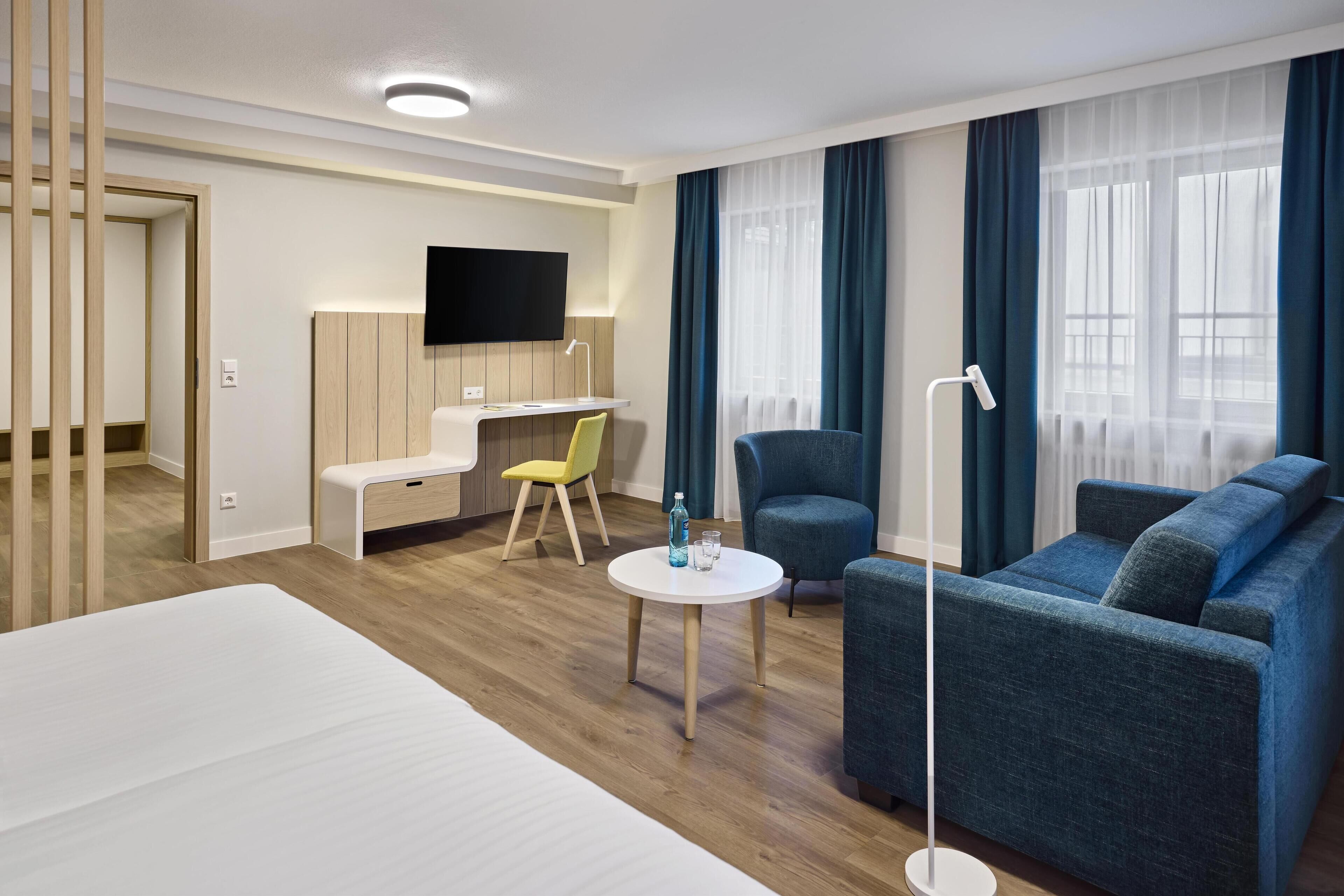 A modern hotel room with a large bed, a desk with a chair, a sofa, a TV mounted on the wall, and a window with curtains.