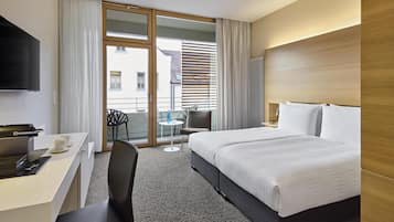 A hotel room with a bed, a desk with a computer, a chair, a small table with a cup, and a view of the outside through a sliding door.