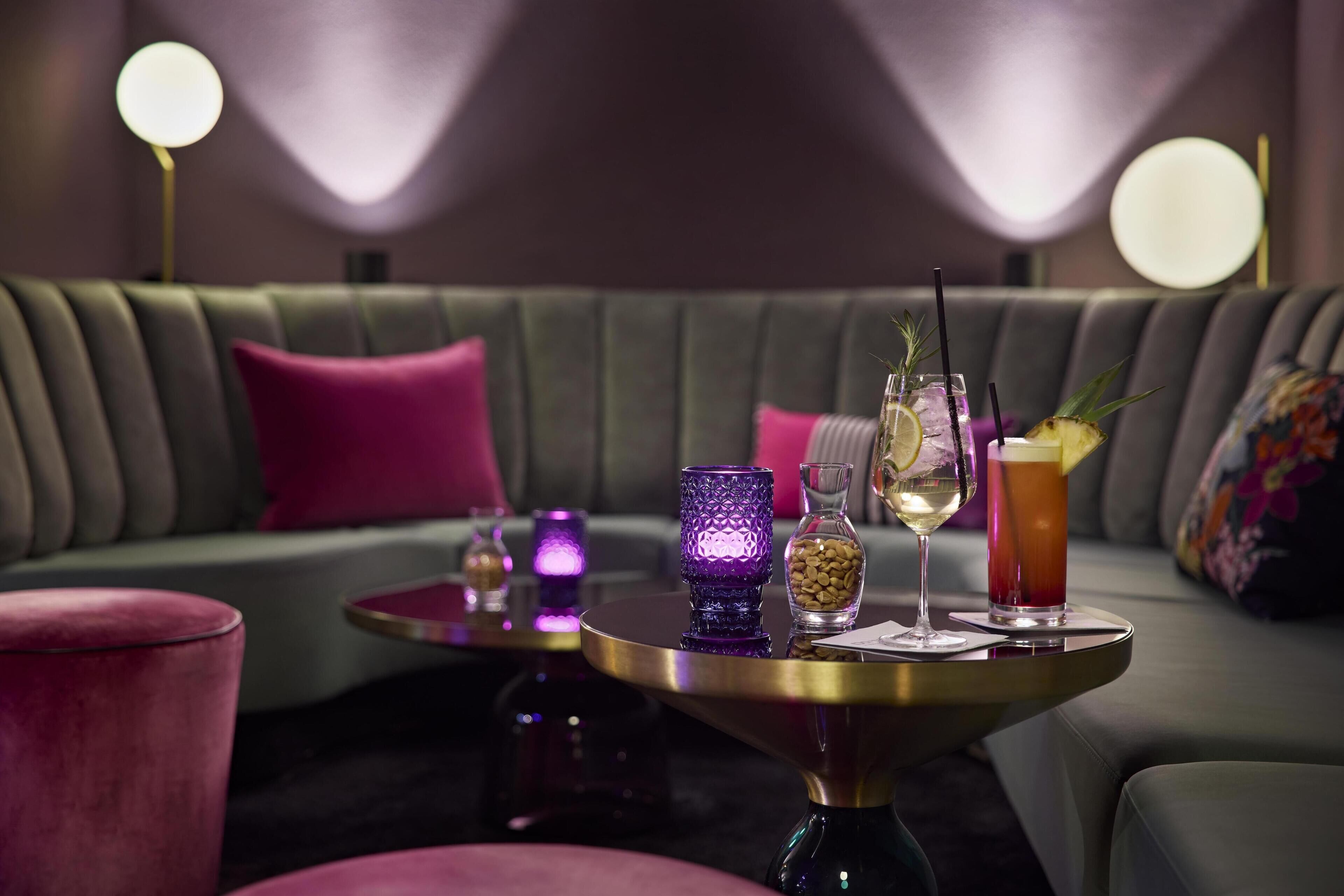 A modern lounge area with a green sofa, a round table with drinks, and decorative lighting.