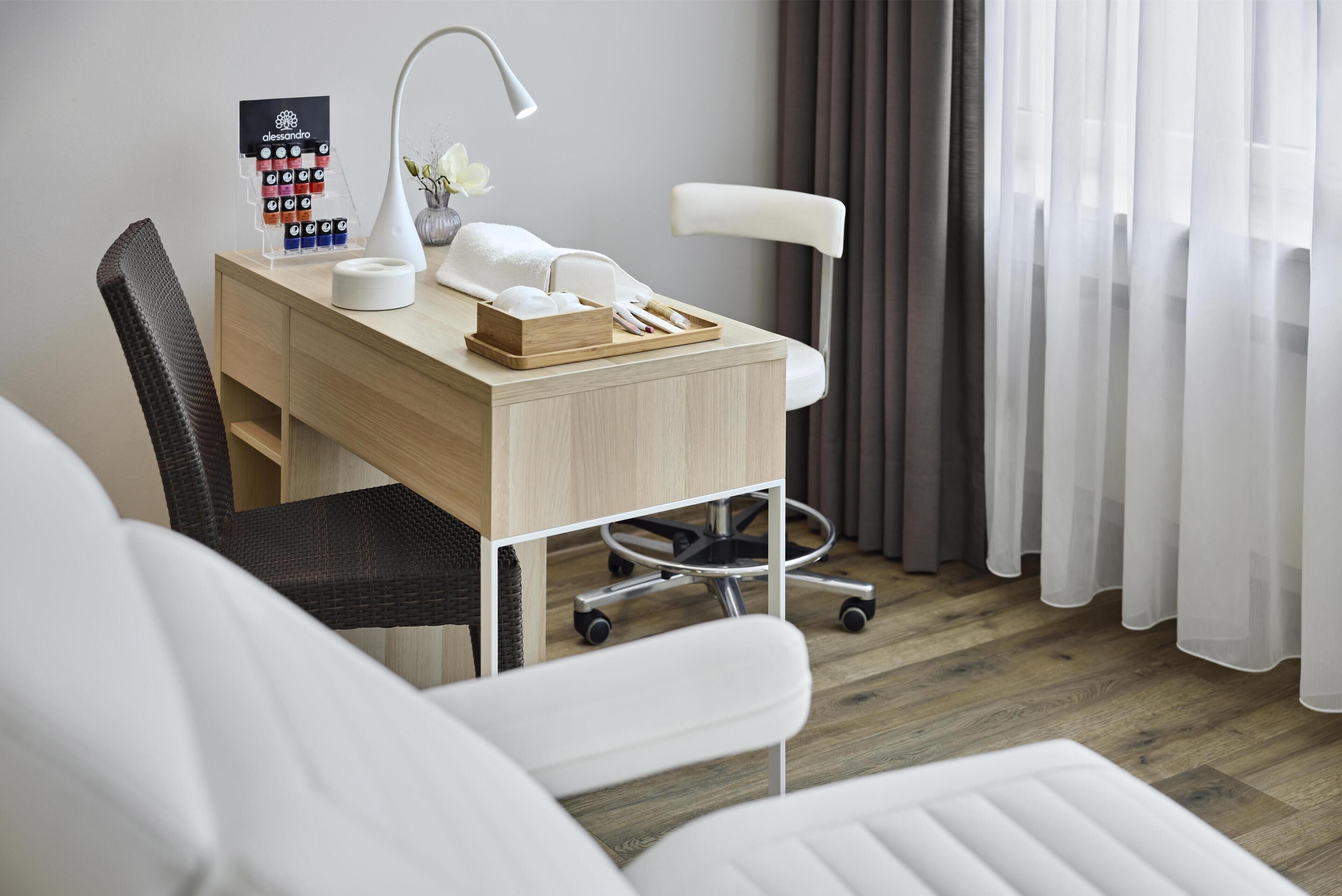 Een moderne hotelkamer met een houten bureau, een witte stoel, een bureaulamp, een kleine vaas met bloemen, en een wielenstoel.