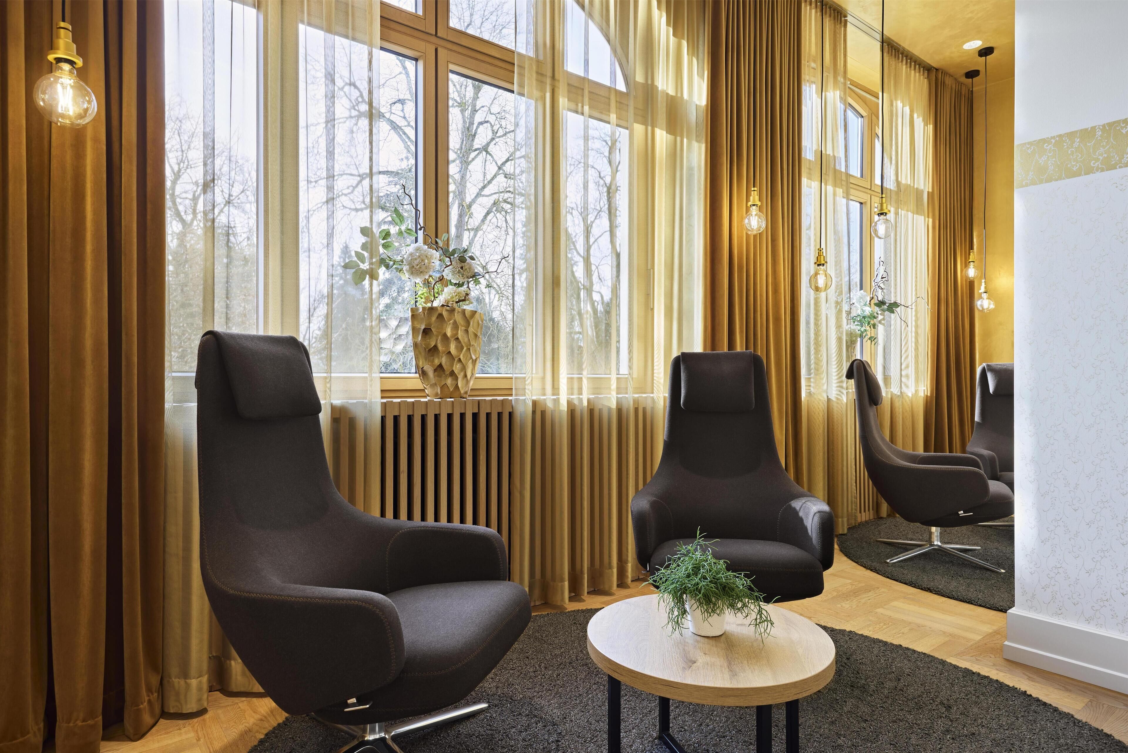 A modern room with three black armchairs, a round wooden table with a plant, and large windows with curtains.
