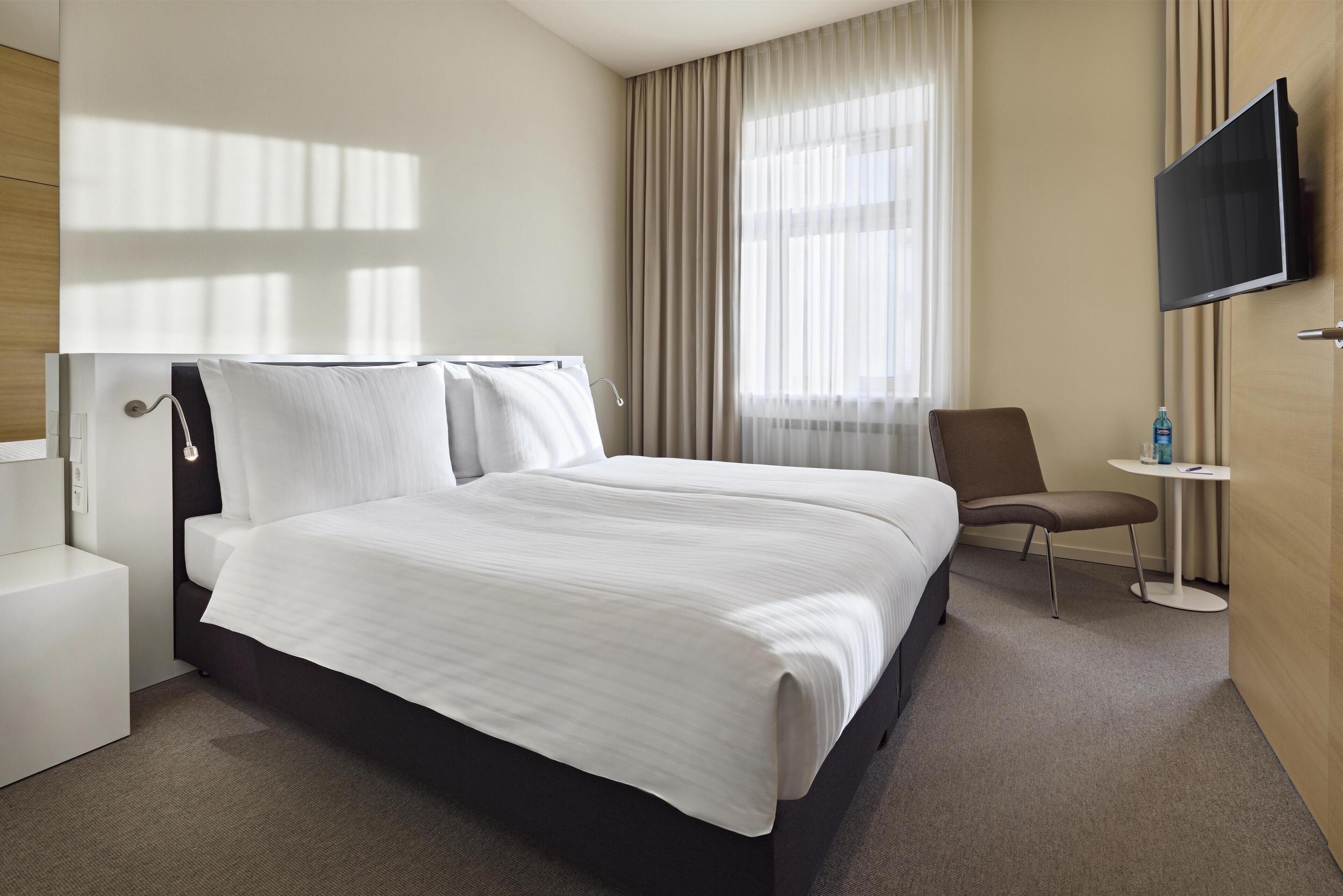 Habitación de hotel con una cama grande, una silla, una mesita, un televisor y una ventana con cortinas.