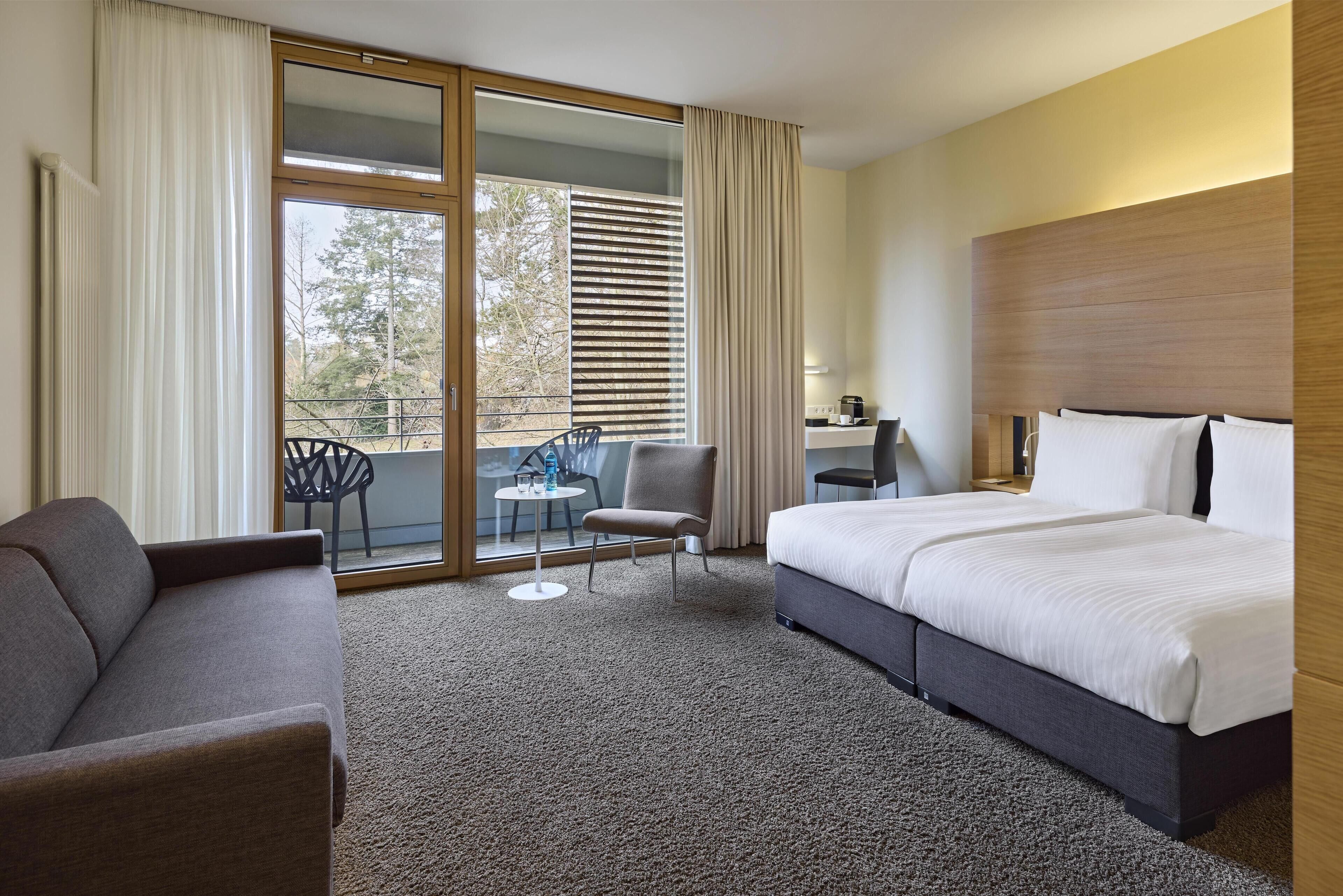 A hotel room with a bed, a sofa, a small table with chairs, a desk with a chair, and a view of the outdoors through a sliding door.
