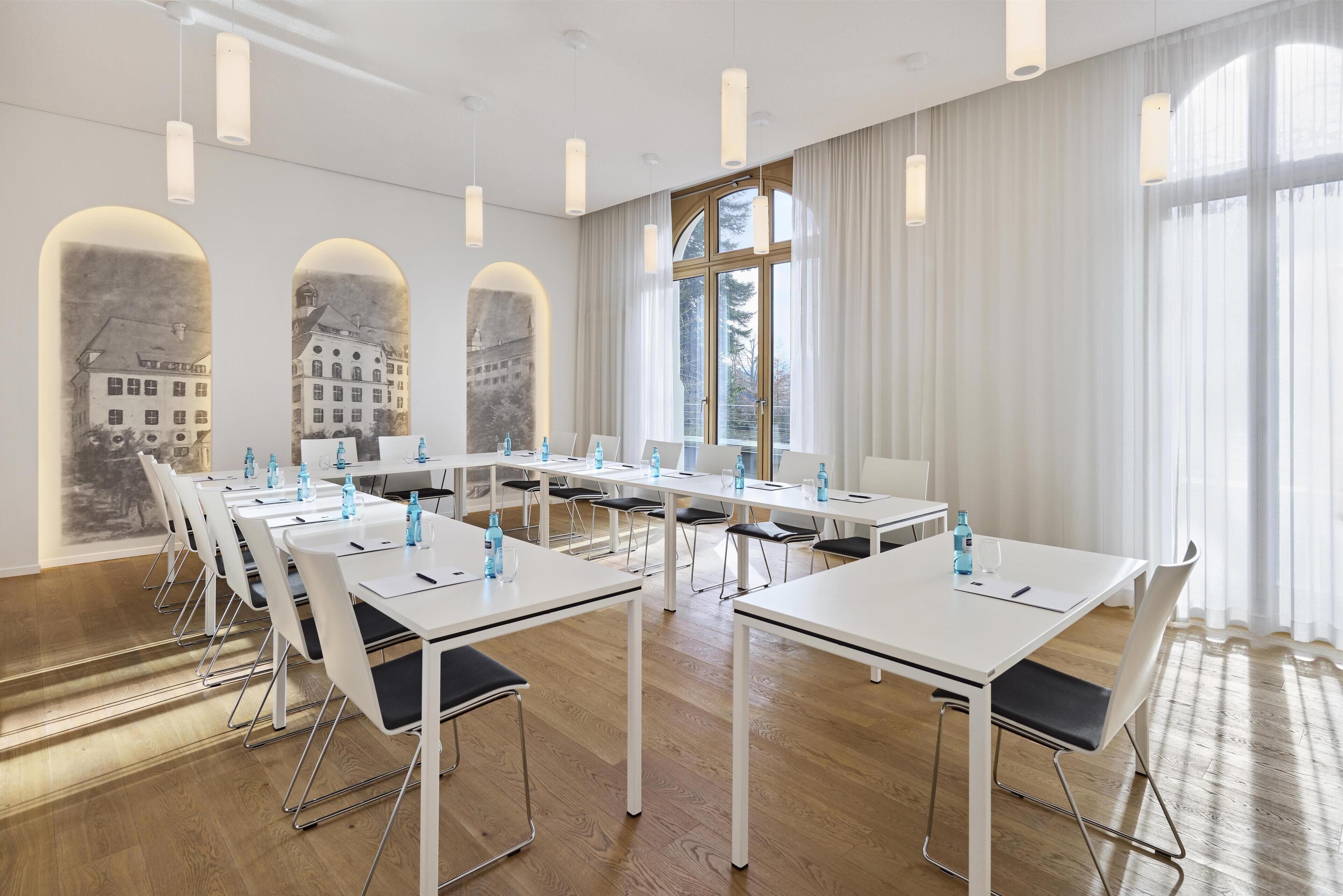 A conference room with white tables and chairs, large windows with sheer curtains, and pendant lighting.