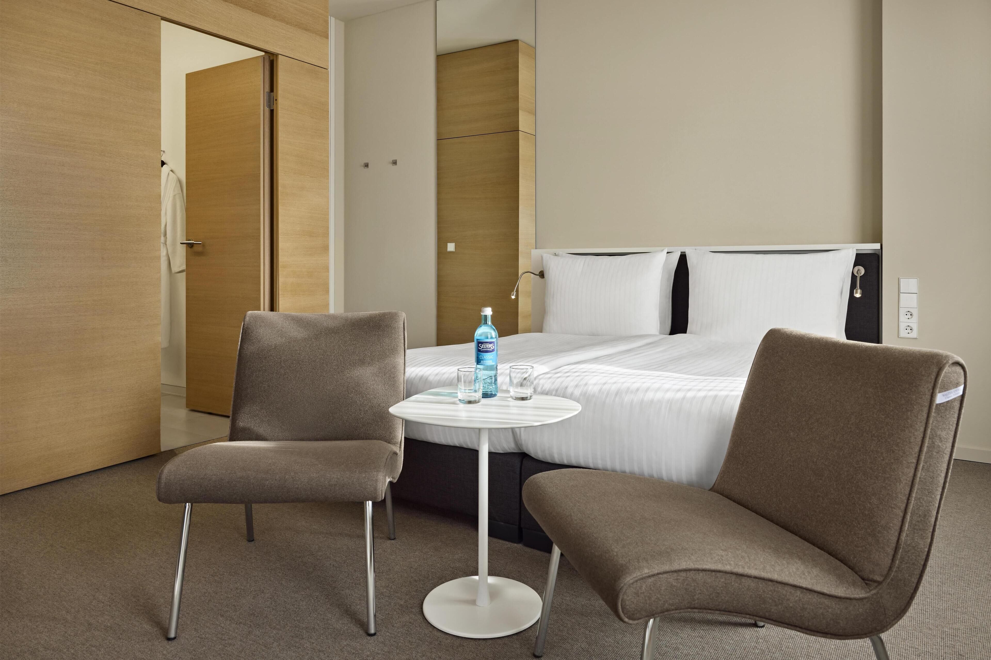 Ett hotellrum med en säng, två fåtöljer, ett litet runt bord och en minibar.