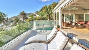 A balcony with lounge chairs, a pool, and a view of a hillside with palm trees. - Clifton Bungalow (Cape Town)