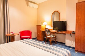 Una habitación de hotel con cama, escritorio con computadora, silla, una silla roja, una mesita con una flor y un espejo.