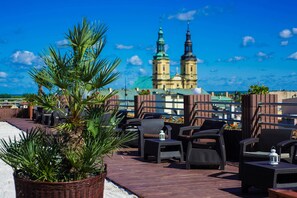 Une terrasse sur le toit, aménagée avec des meubles en osier, un pot de fleurs et offrant une vue sur un bâtiment historique doté de deux flèches.