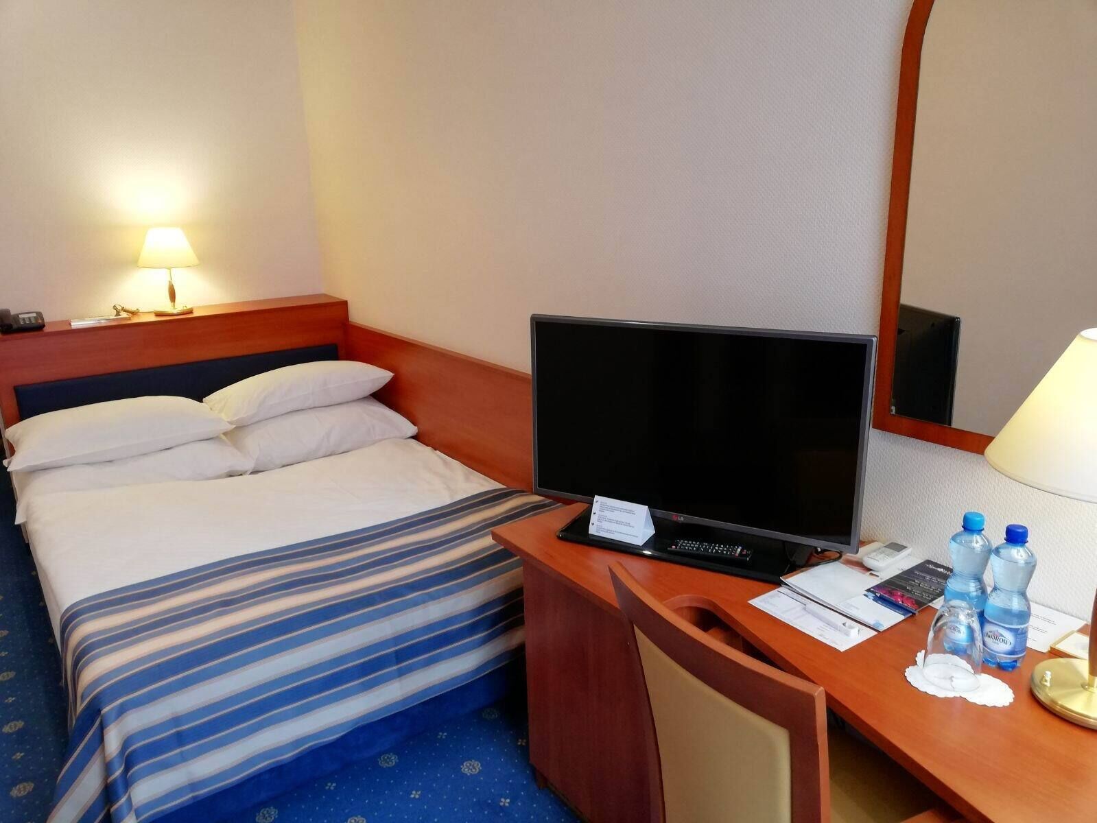 A hotel room with a bed, a desk with a computer, a chair, and a lamp.