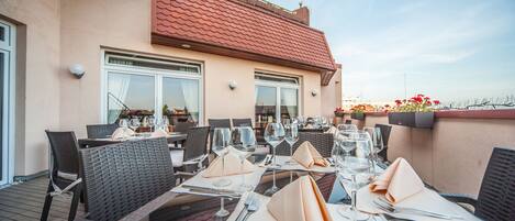An outdoor dining area with tables set for two, featuring wicker chairs and glassware.