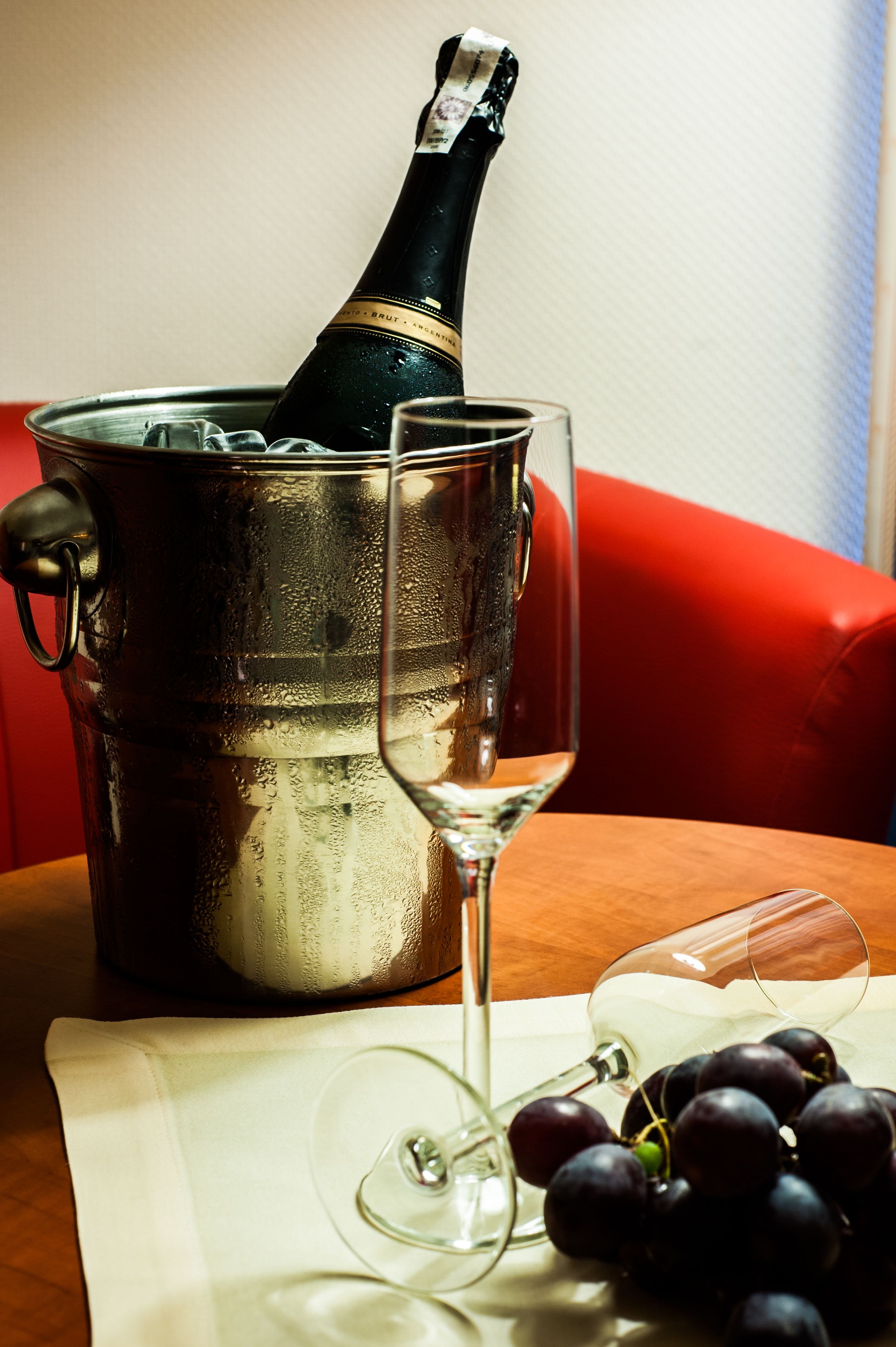 A bottle of champagne in a metal bucket, two wine glasses, and a bunch of grapes on a table.