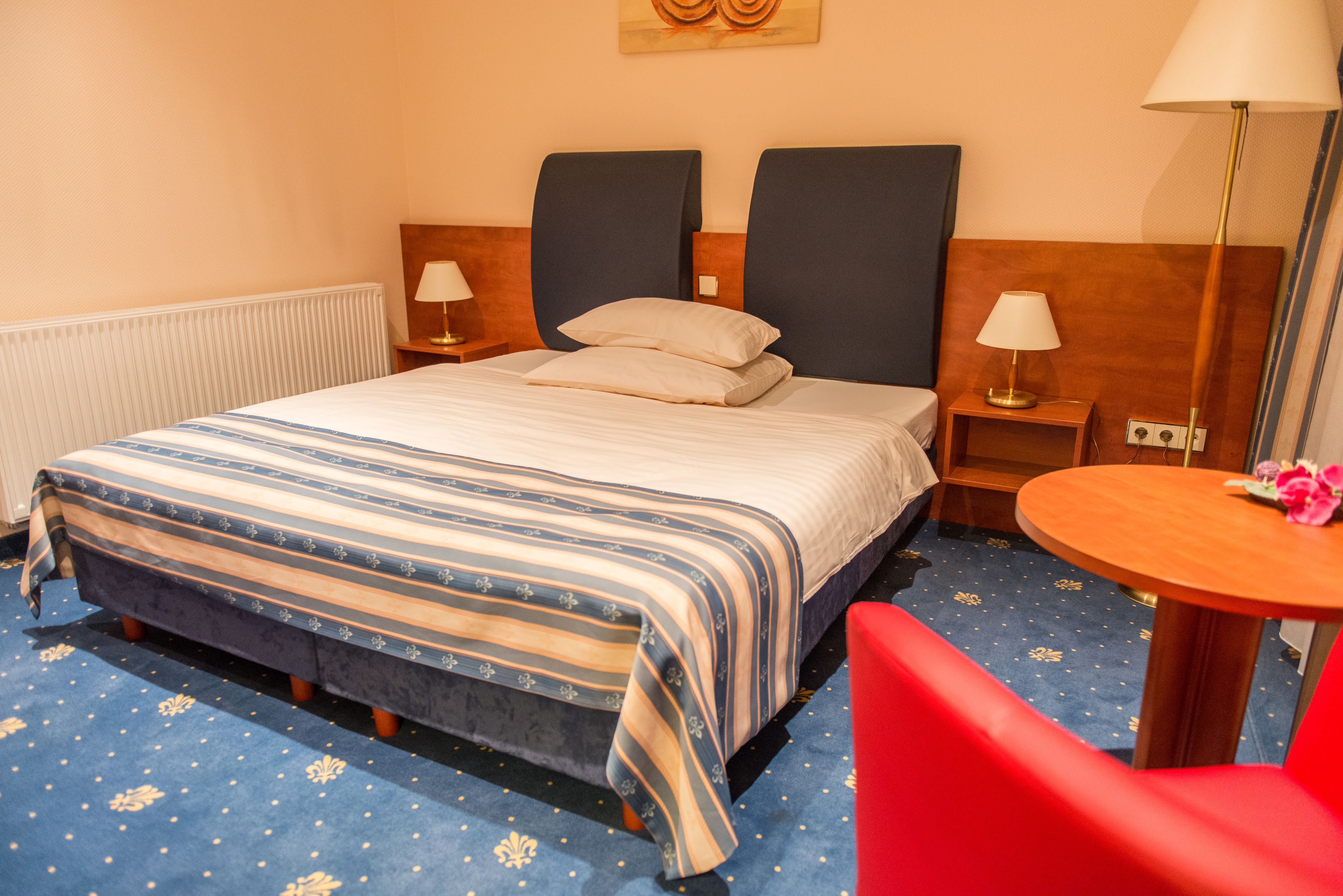 A hotel room with a large bed, two bedside tables with lamps, a small round table with a flower, and a red chair.