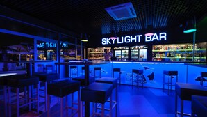 A bar with a neon sign reading "SKYLIGHT BAR" and a bartender. There are several bar stools and a counter with a variety of bottles and glasses.