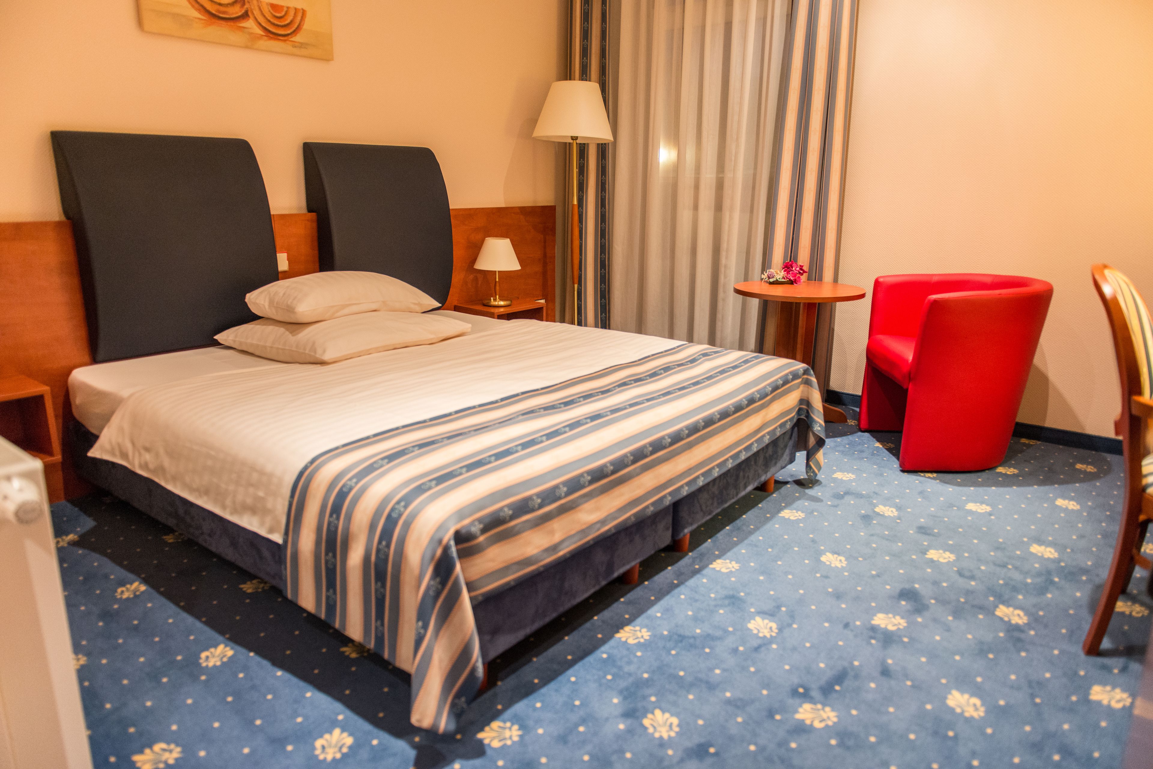 Una habitación de hotel con una cama, una silla roja, una mesita y una lámpara.