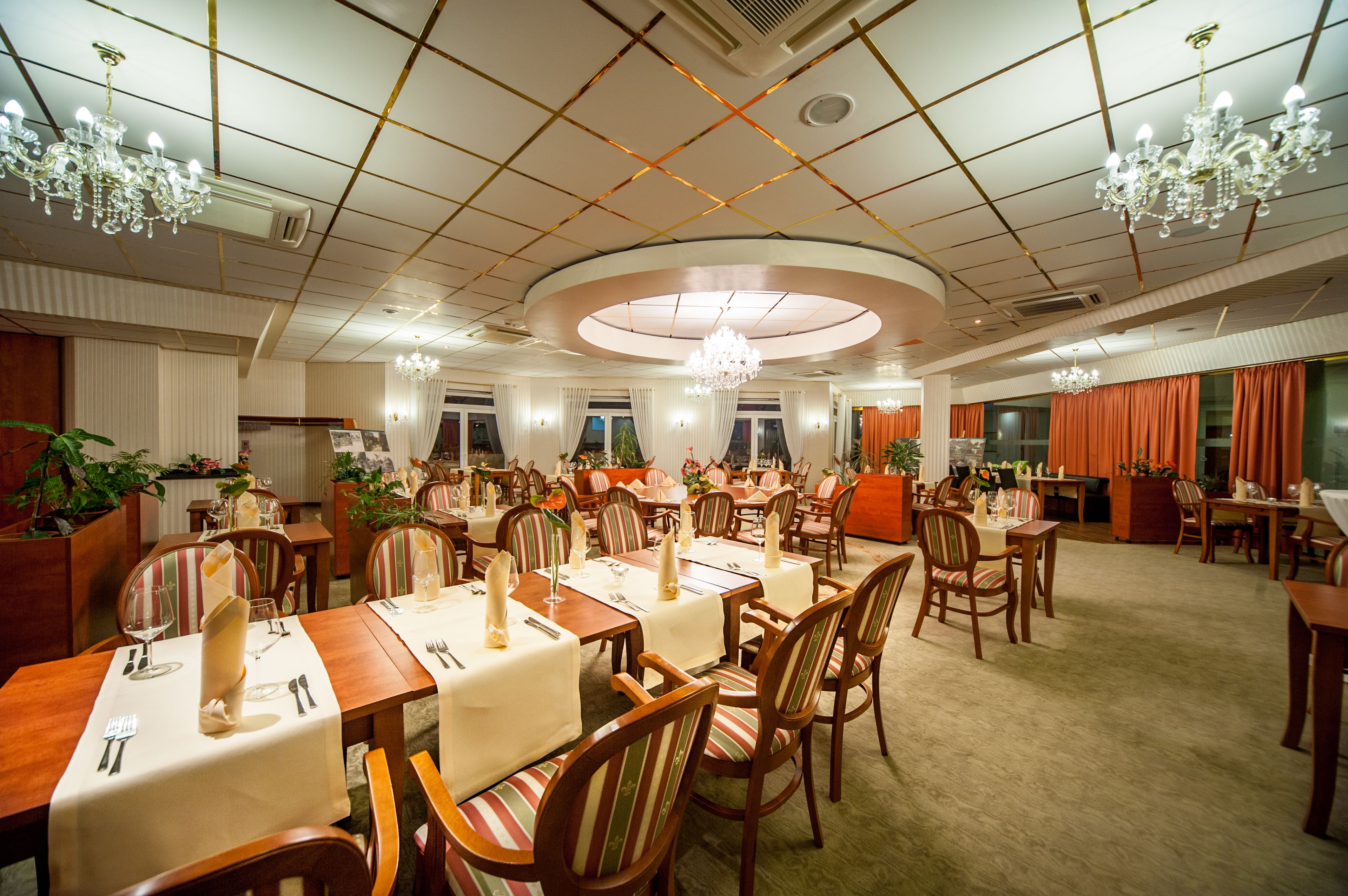 A spacious dining area with multiple tables and chairs, chandeliers, and potted plants.