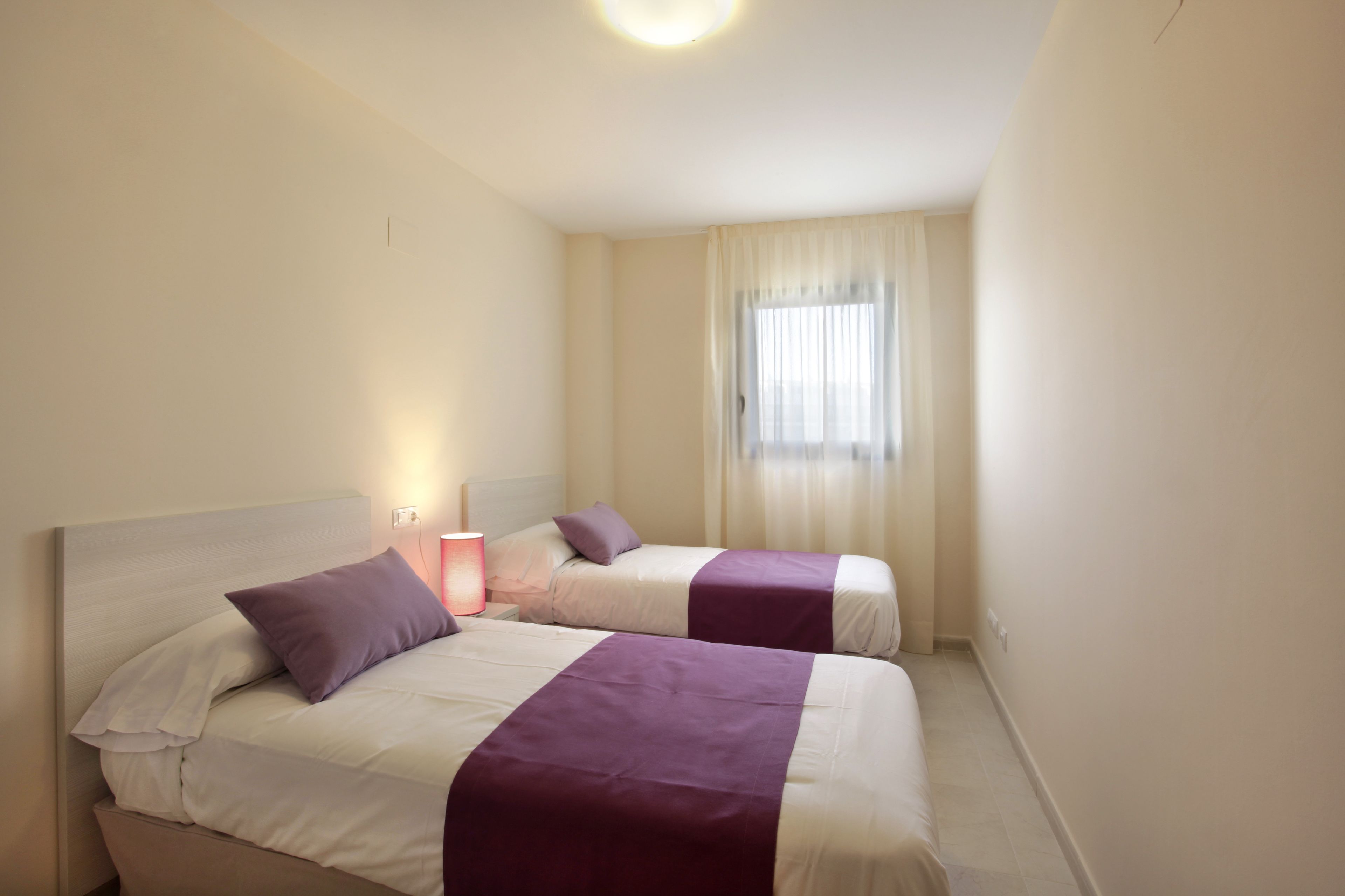 Habitación de hotel con dos camas, cada una con una colcha morada y ropa de cama blanca. Hay una mesita de noche con una lámpara, una ventana con cortinas traslúcidas y una luz en el techo.