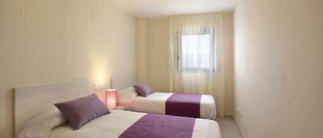Habitación de hotel con dos camas, cada una con una colcha morada y ropa de cama blanca. Hay una mesita de noche con una lámpara, una ventana con cortinas traslúcidas y una luz en el techo.