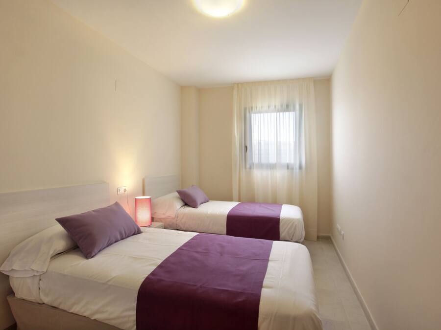 Une chambre d’hôtel avec deux lits, chacun recouvert d’une couvre-lit violette et doté de draps blancs. On y trouve une table de chevet avec une lampe, une fenêtre avec des rideaux légers et un luminaire au plafond.