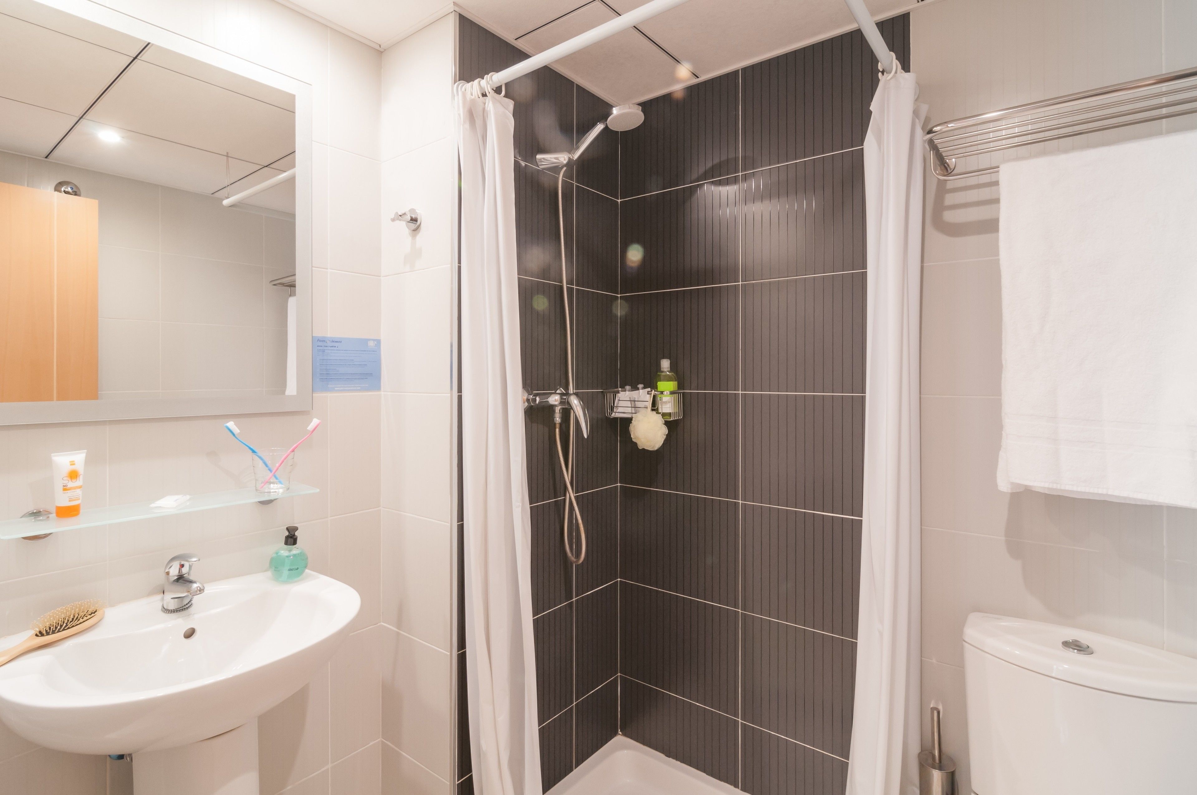 A modern bathroom with a white sink, a glass shelf with toiletries, a shower with a curtain, and a toilet.