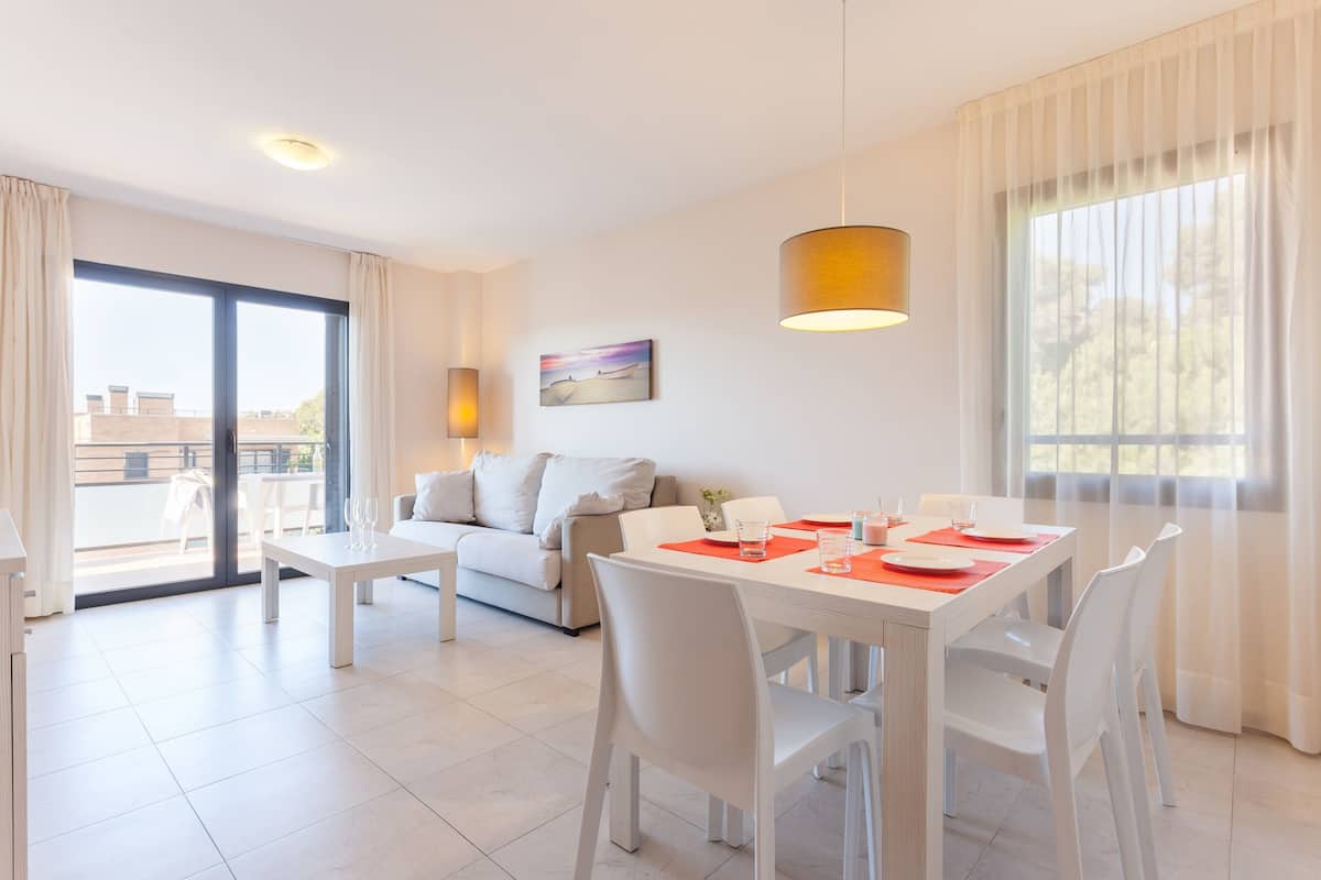 A modern living room with a dining area, featuring a sofa, a dining table with chairs, a pendant light, and a large window with curtains.