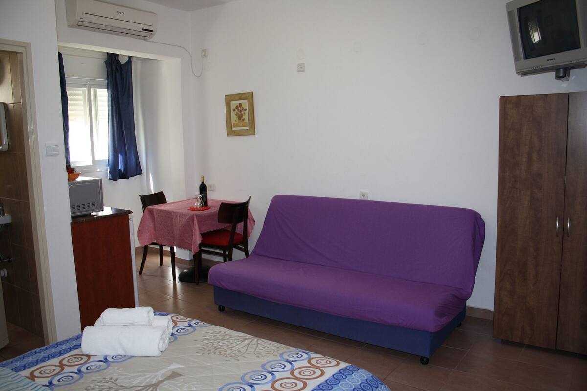 A room with a bed, purple sofa, wooden wardrobe, and a small dining area with a table and chairs.
