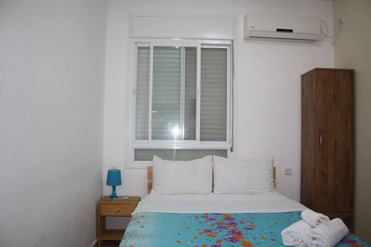 A bedroom with a bed, a nightstand, a lamp, a wardrobe, and a window with blinds.