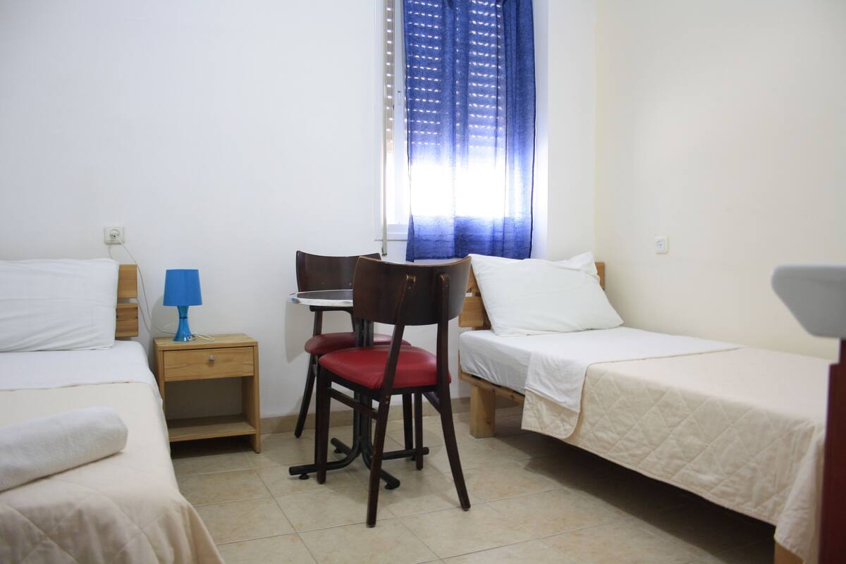 A small, clean room with two single beds, a wooden nightstand with a lamp, a chair, and a desk.