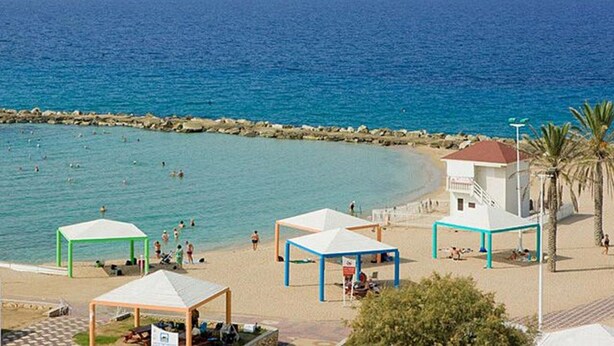 A beach with clear blue water, a rocky breakwater, and several beach huts. - Ga'aton Motel (Nahariya)