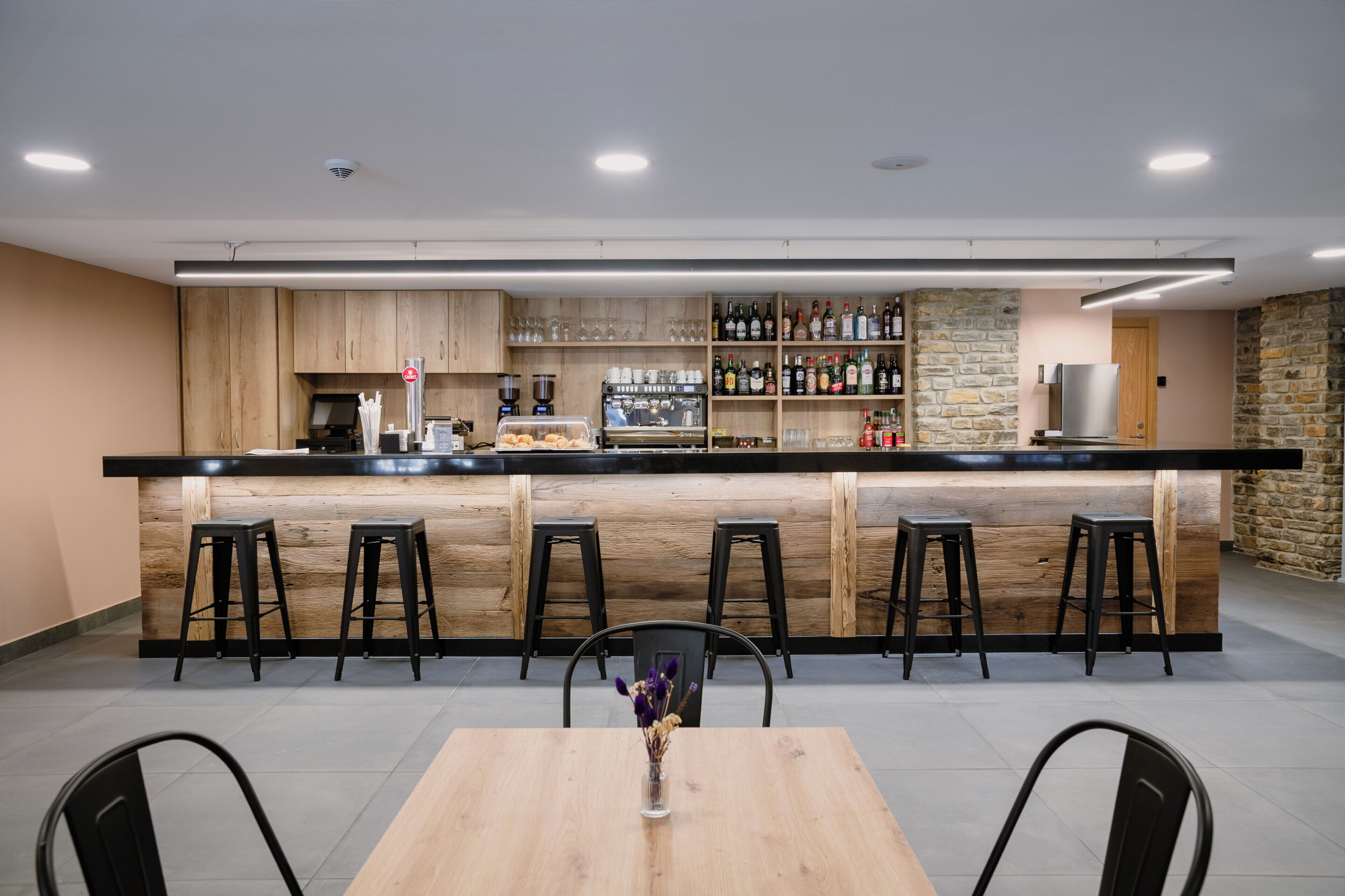 A modern bar area with a wooden counter, bar stools, a coffee machine, and a variety of bottles.