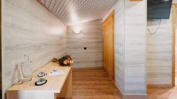 Habitación de hotel con suelo de madera, un mostrador de recepción con una botella y un vaso de vidrio, un ramo de flores y un armario de madera.