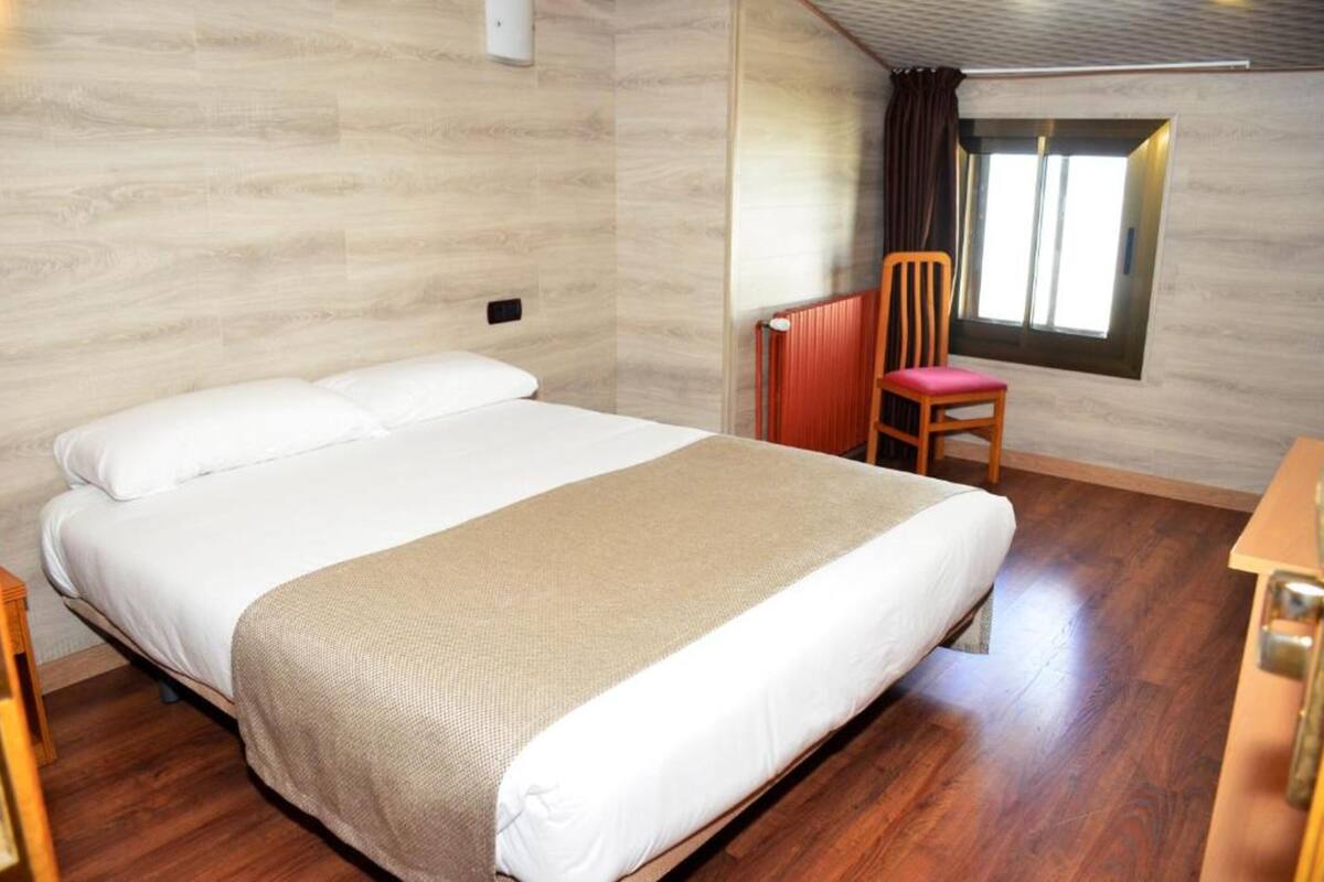 Une chambre à coucher avec un grand lit, une chaise en bois, une fenêtre avec des rideaux et un radiateur.