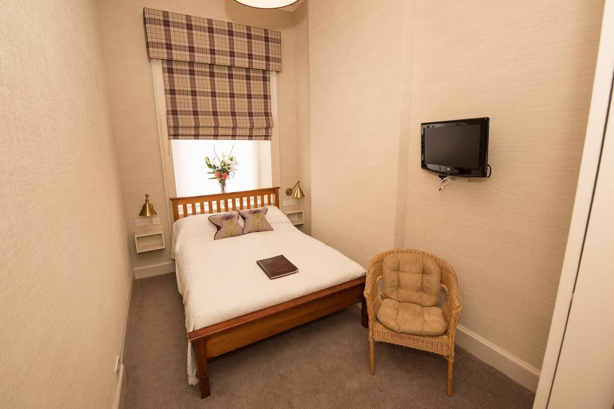 A bedroom with a bed, a chair, a TV, a window with a plaid valance, and a bedside table with lamps.