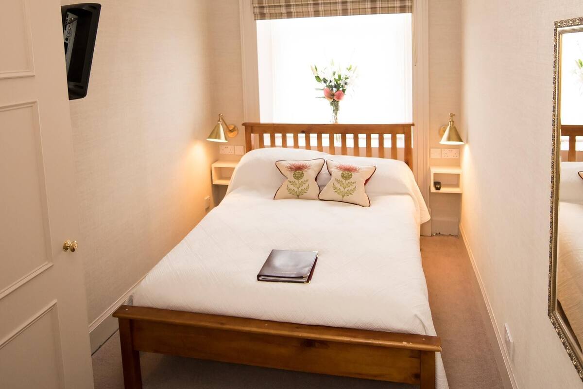 A neatly arranged bedroom with a wooden bed, bedside lamps, a window with plaid curtains, and a small bedside table with a lamp.