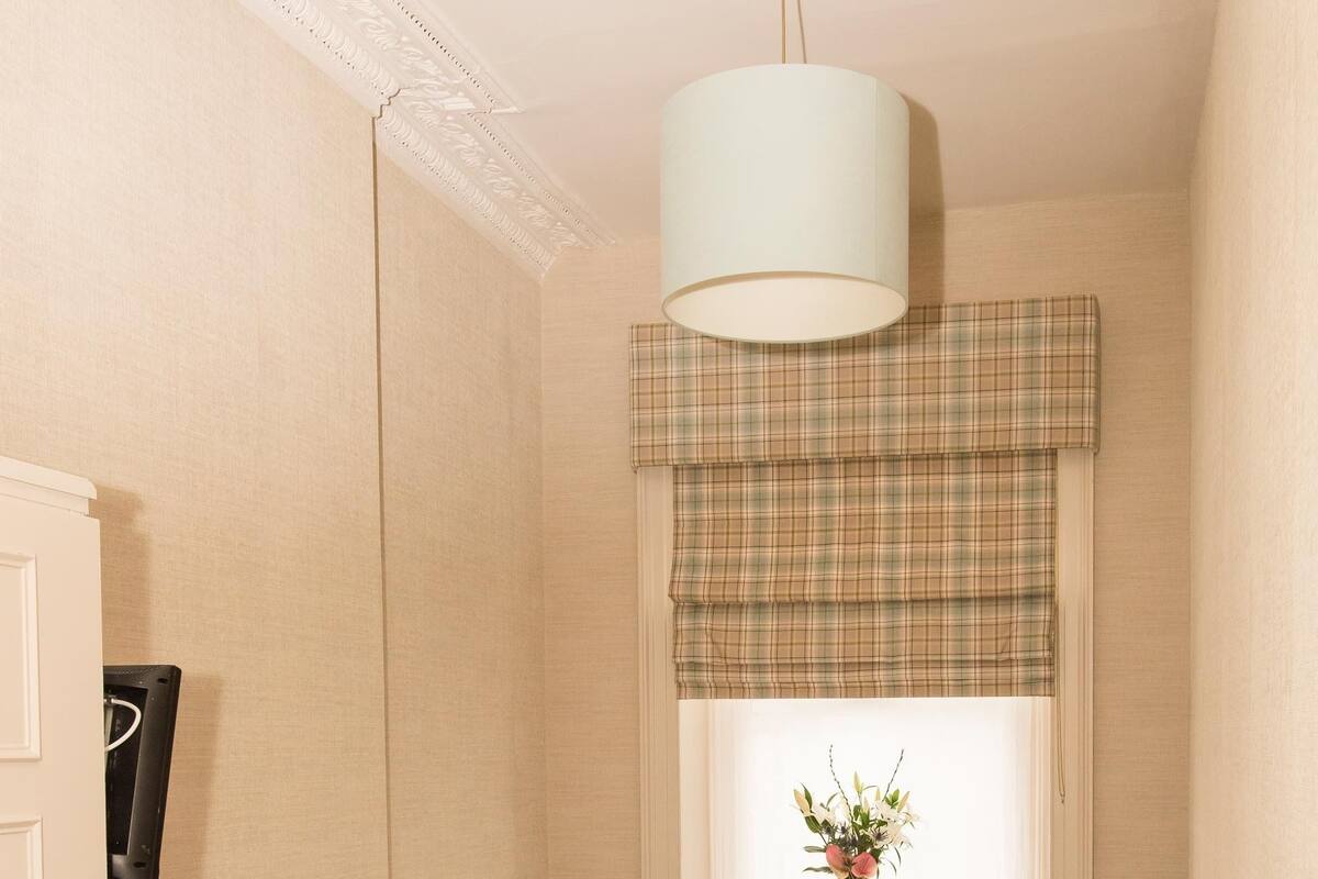 A bedroom with two single beds, a wicker chair, a bedside table with a lamp, and a window with a plaid valance.