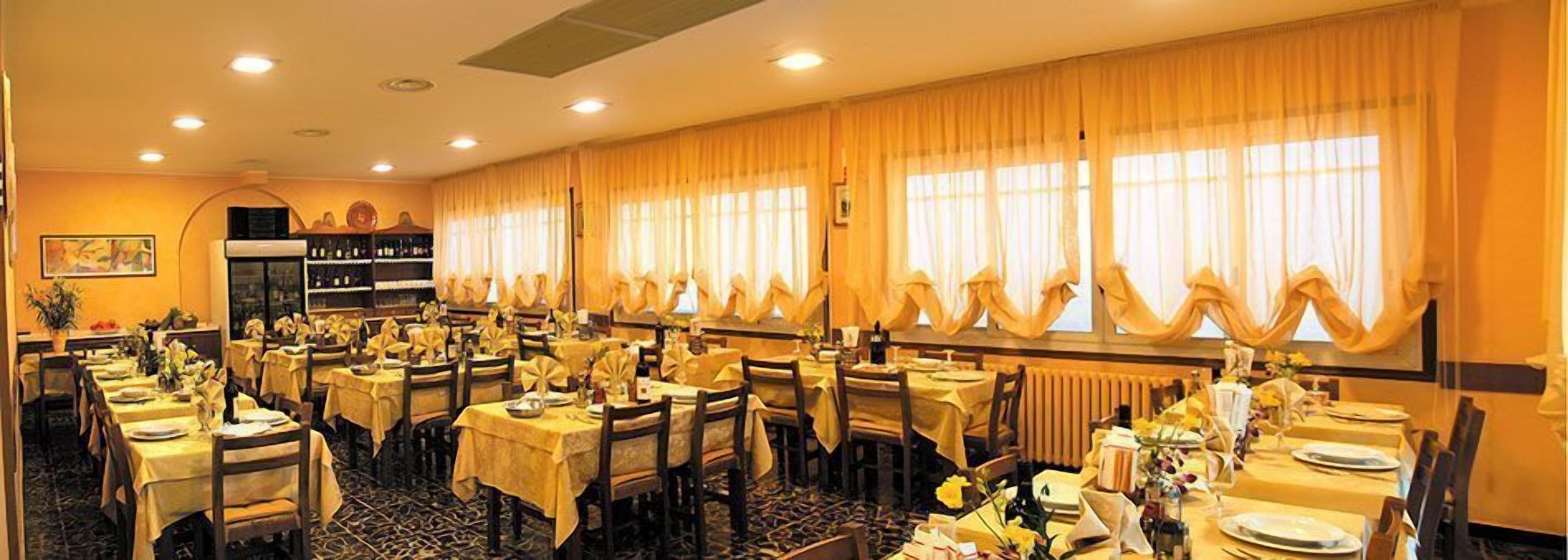 Een hotelrestaurant met meerdere gedekte tafels, grote ramen met gordijnen en een zichtbare wijnkast.