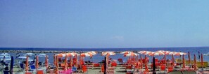 A beach with rows of beach chairs and umbrellas, a rocky shoreline, and the ocean in the background. - Hotel Candido (Diano Marina)