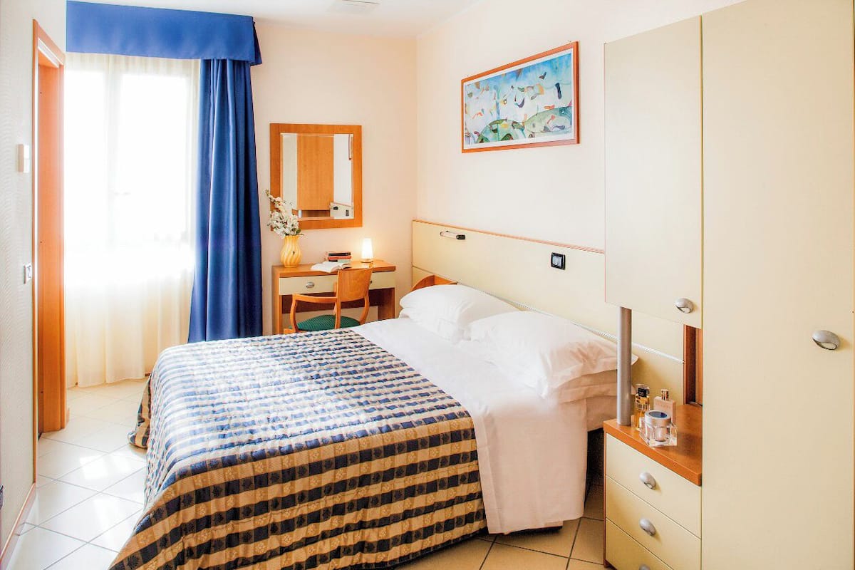 Een hotelkamer met een bed, een bureau, een stoel, een kast, een spiegel en een schilderij aan de muur.